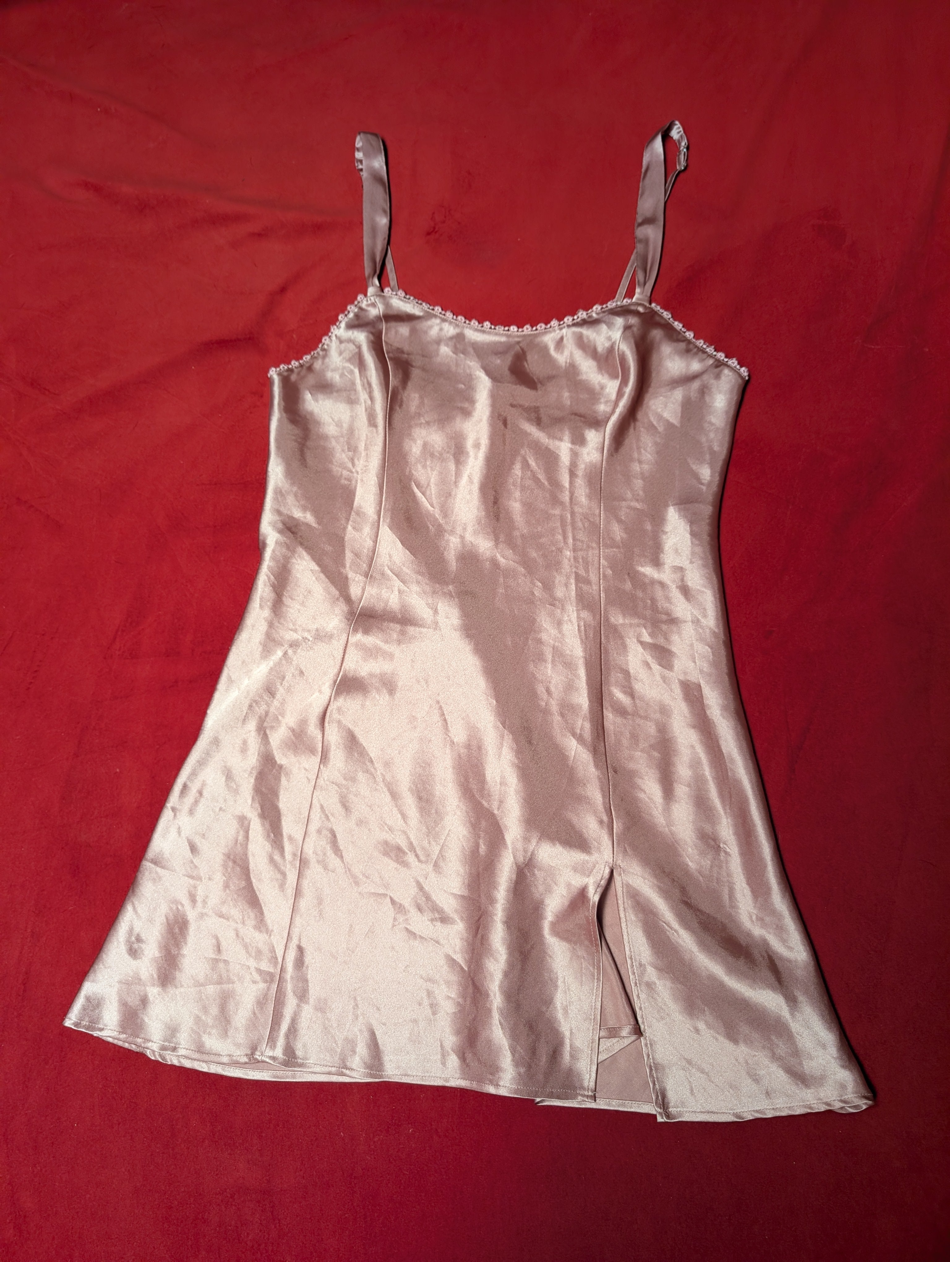 A light pink satin slip dress with thin shoulder straps and a scalloped neckline, laid flat on a red fabric background.