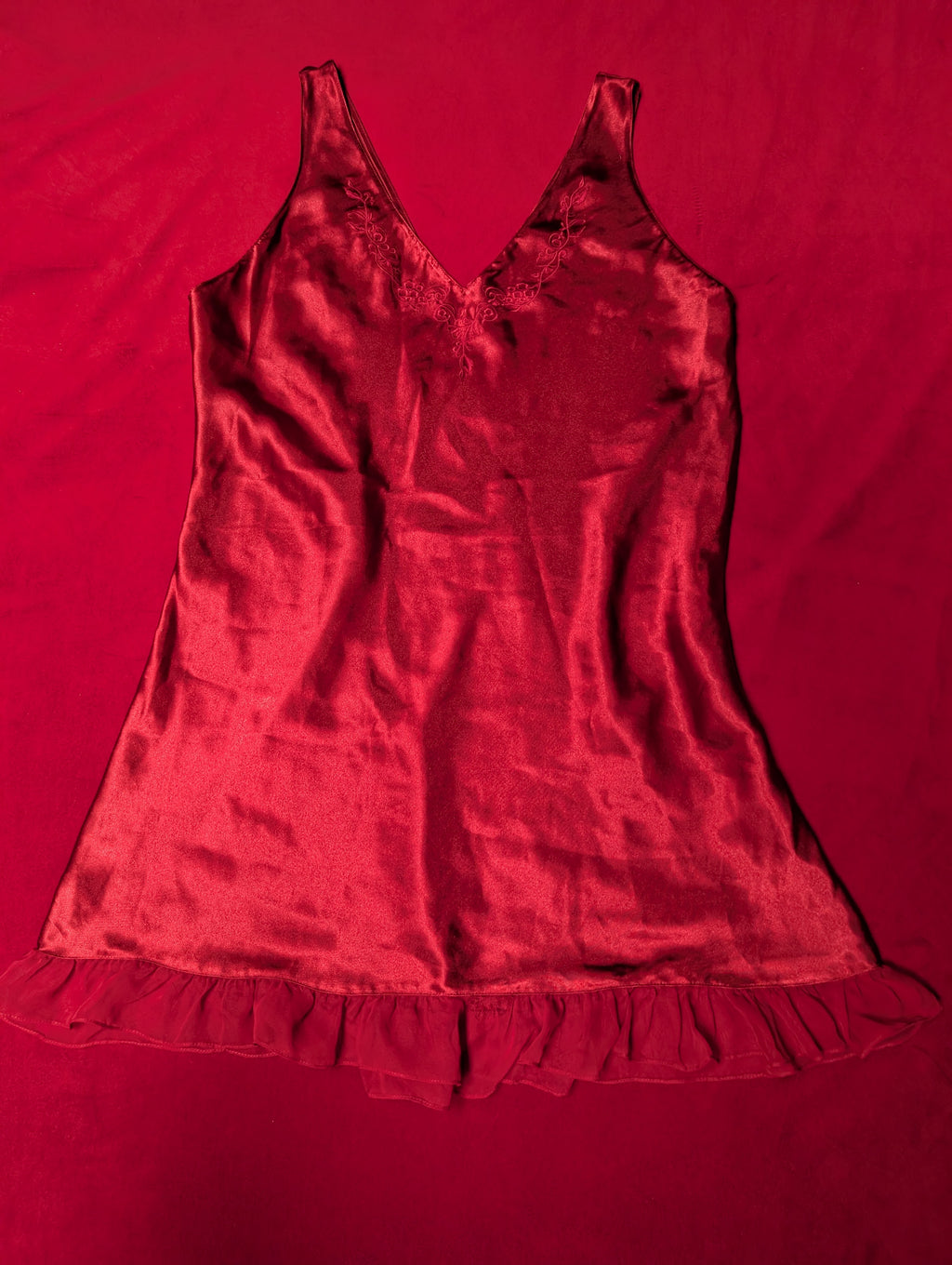 A red satin camisole chemise with a ruffled hem, laid flat on a matching red fabric background.