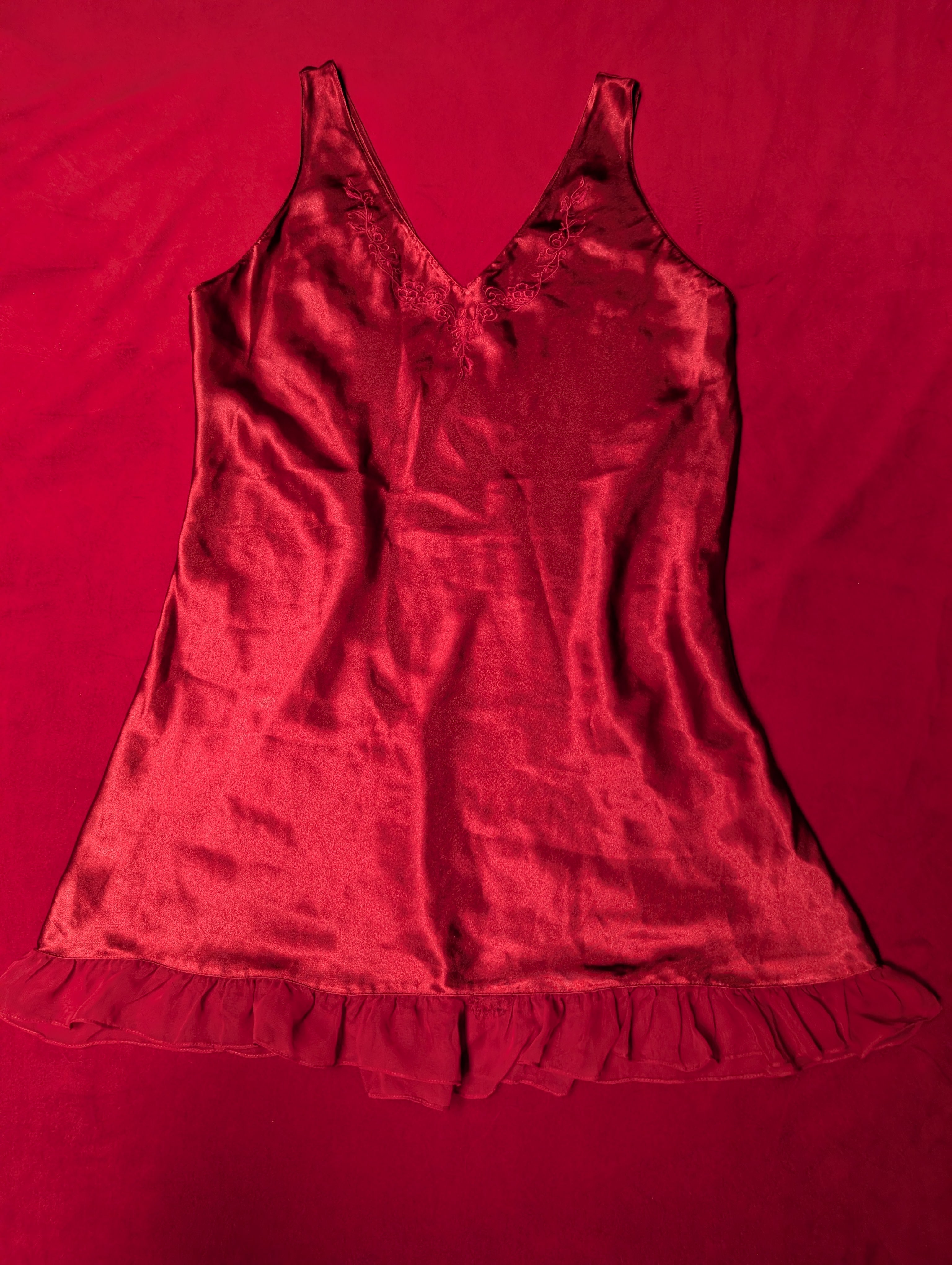 A red satin camisole chemise with a ruffled hem, laid flat on a matching red fabric background.