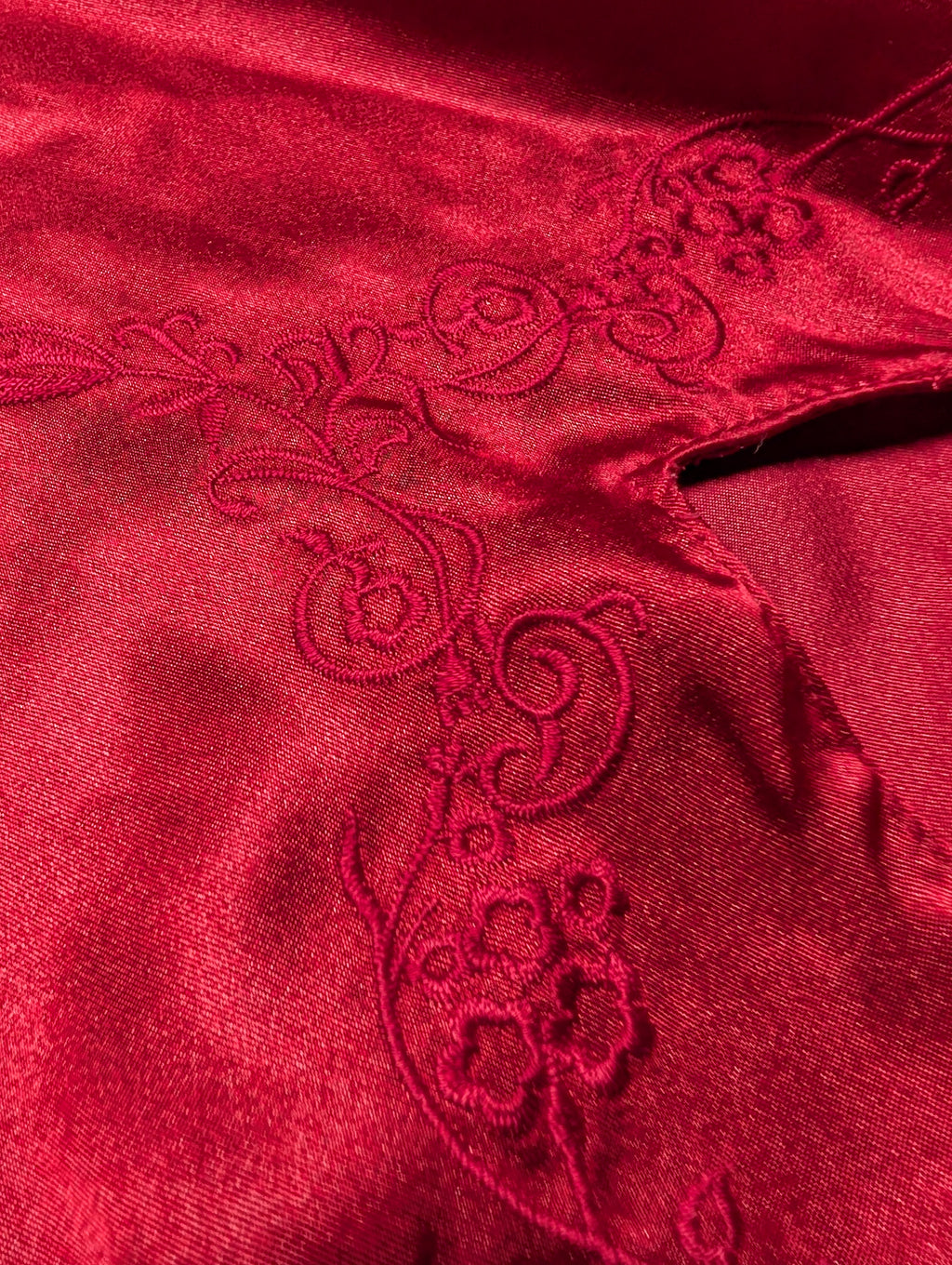 A close-up of a rich red satin fabric with matching red embroidered floral and scrollwork details.