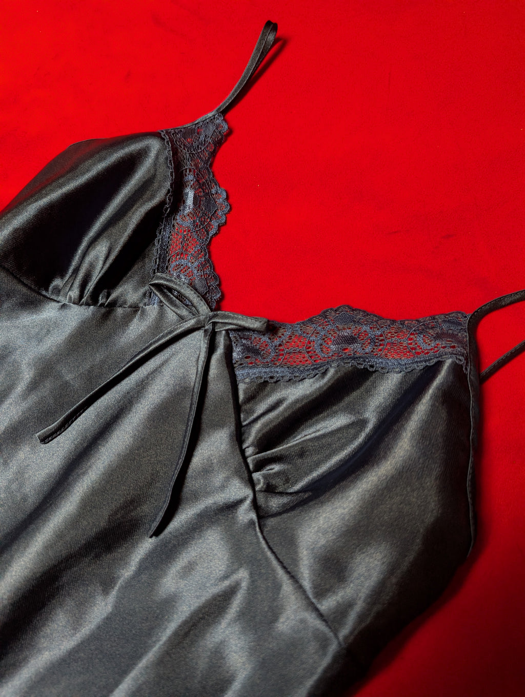 A close-up of a dark navy blue satin slip dress or lingerie top with thin straps and a lace neckline, laid flat on a bright red background.