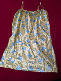 A butter yellow satin slip dress with a blue and green floral pattern, featuring thin spaghetti straps, laid flat against a dark red background.