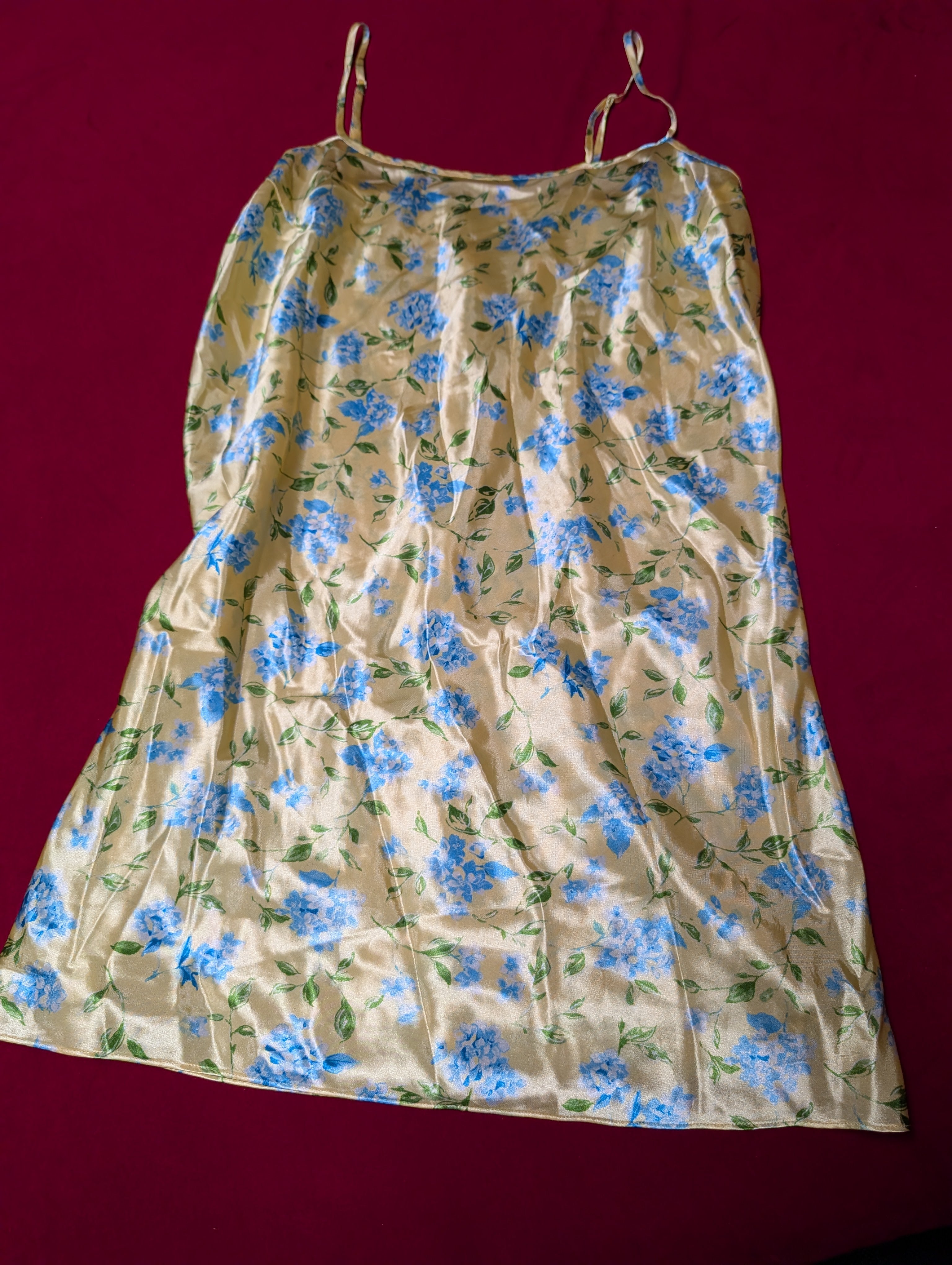 A butter yellow satin slip dress with a blue and green floral pattern, featuring thin spaghetti straps, laid flat against a dark red background.