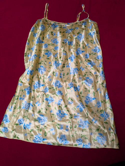 A butter yellow satin slip dress with a blue and green floral pattern, featuring thin spaghetti straps, laid flat against a dark red background.