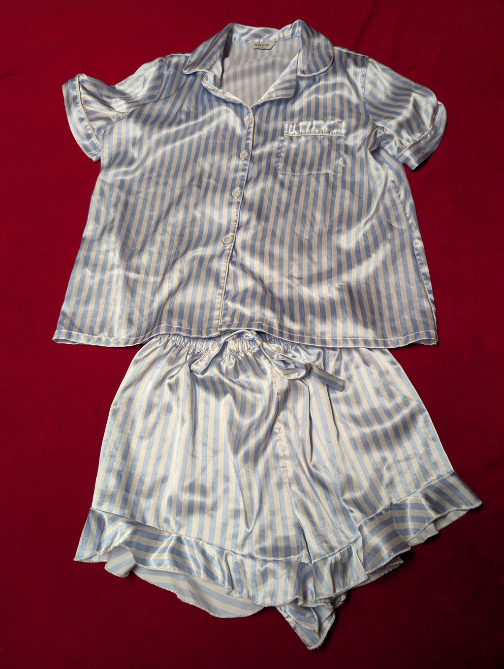 A two-piece women's pajama set made of shiny satin-like fabric with vertical blue and white stripes, laid flat on a red background. The set includes a short-sleeve button-front top with a collar and chest pocket, and matching ruffled shorts with an elastic drawstring waist.