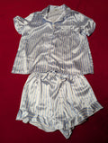 A two-piece women's pajama set made of shiny satin-like fabric with vertical blue and white stripes, laid flat on a red background. The set includes a short-sleeve button-front top with a collar and chest pocket, and matching ruffled shorts with an elastic drawstring waist.