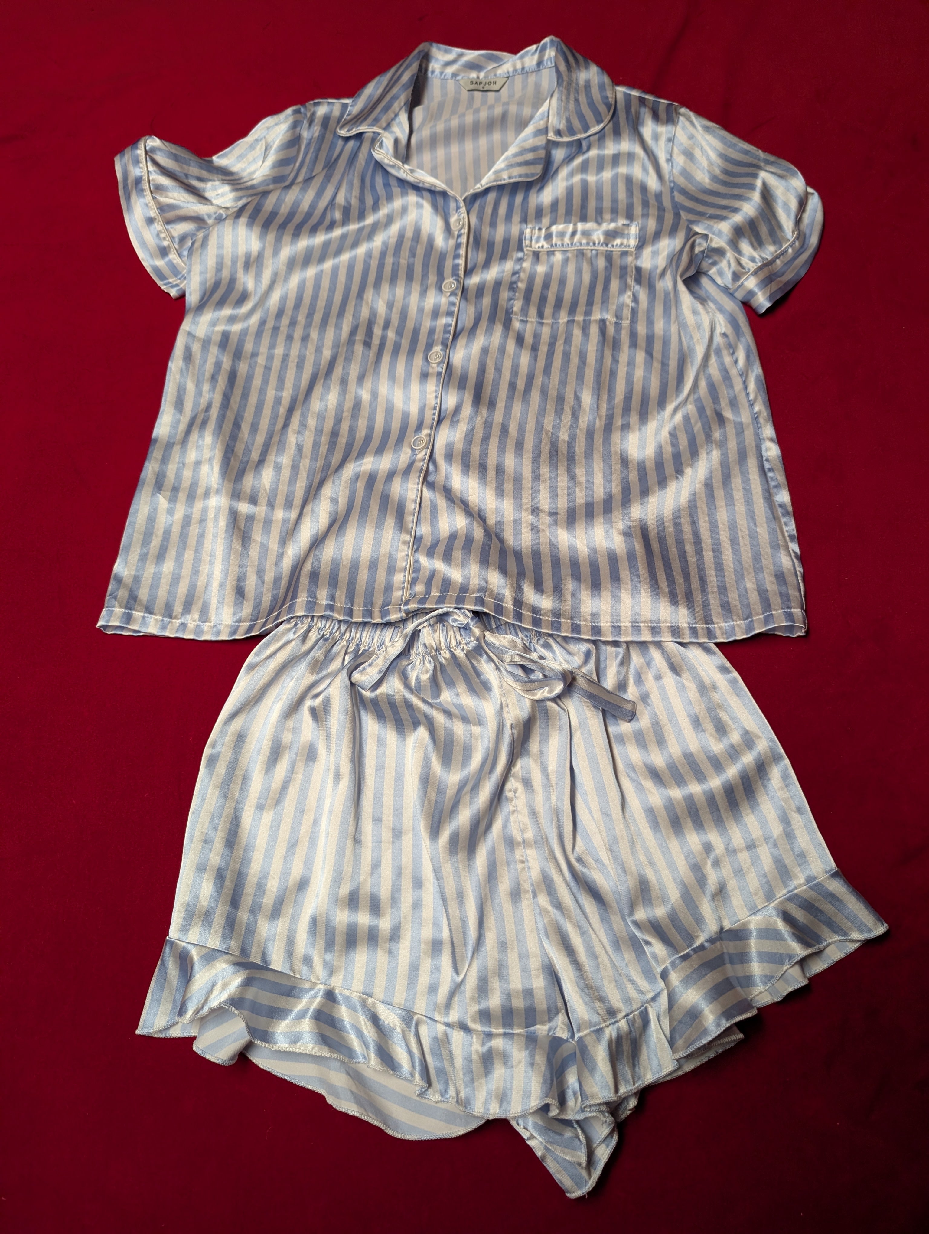 A two-piece women's pajama set made of shiny satin-like fabric with vertical blue and white stripes, laid flat on a red background. The set includes a short-sleeve button-front top with a collar and chest pocket, and matching ruffled shorts with an elastic drawstring waist.