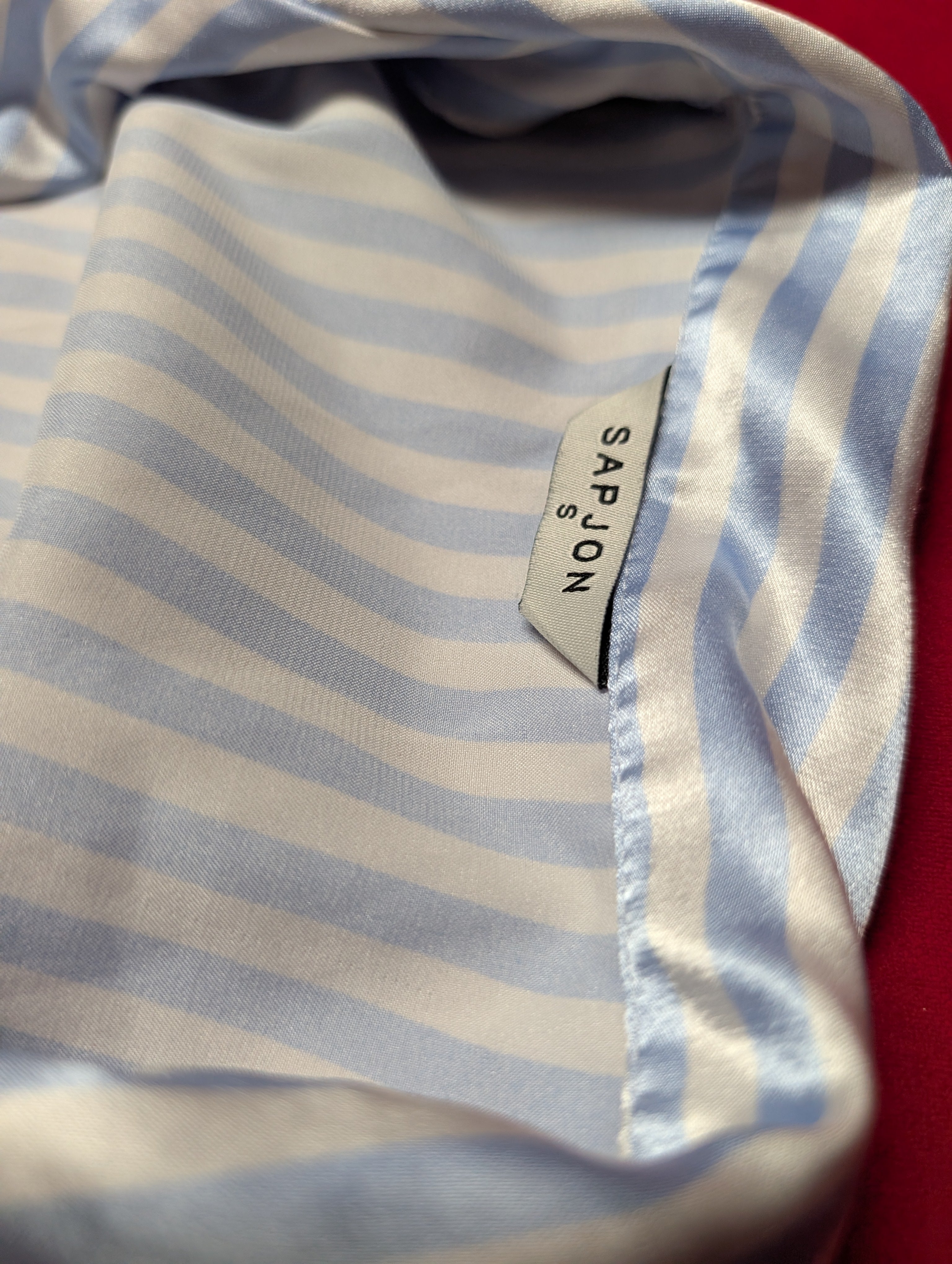 A close-up of a light blue and white striped fabric item, likely a scarf or shawl, featuring a visible SAPJON brand label.