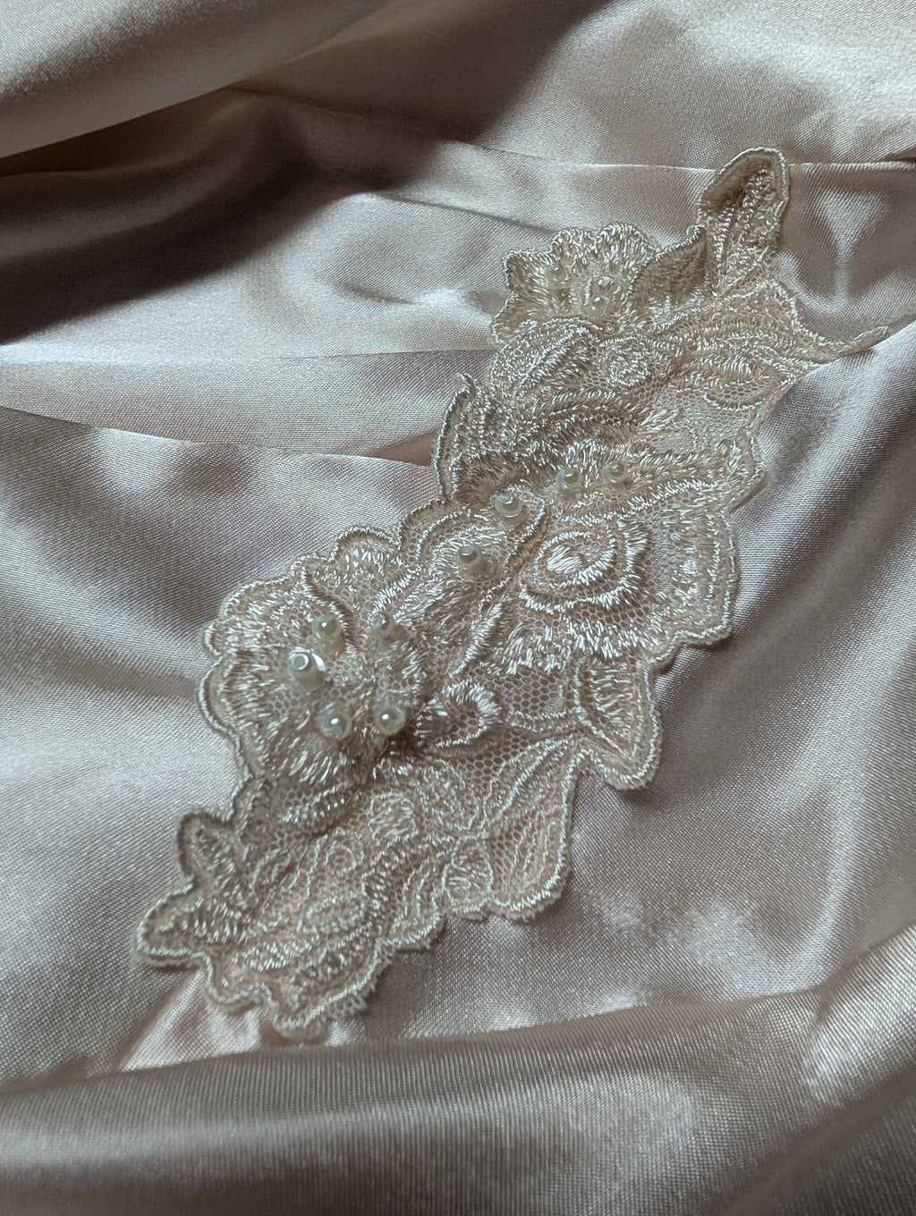 A close-up of a cream-colored floral lace applique with pearl embellishments, laid flat on a draped champagne satin fabric background.