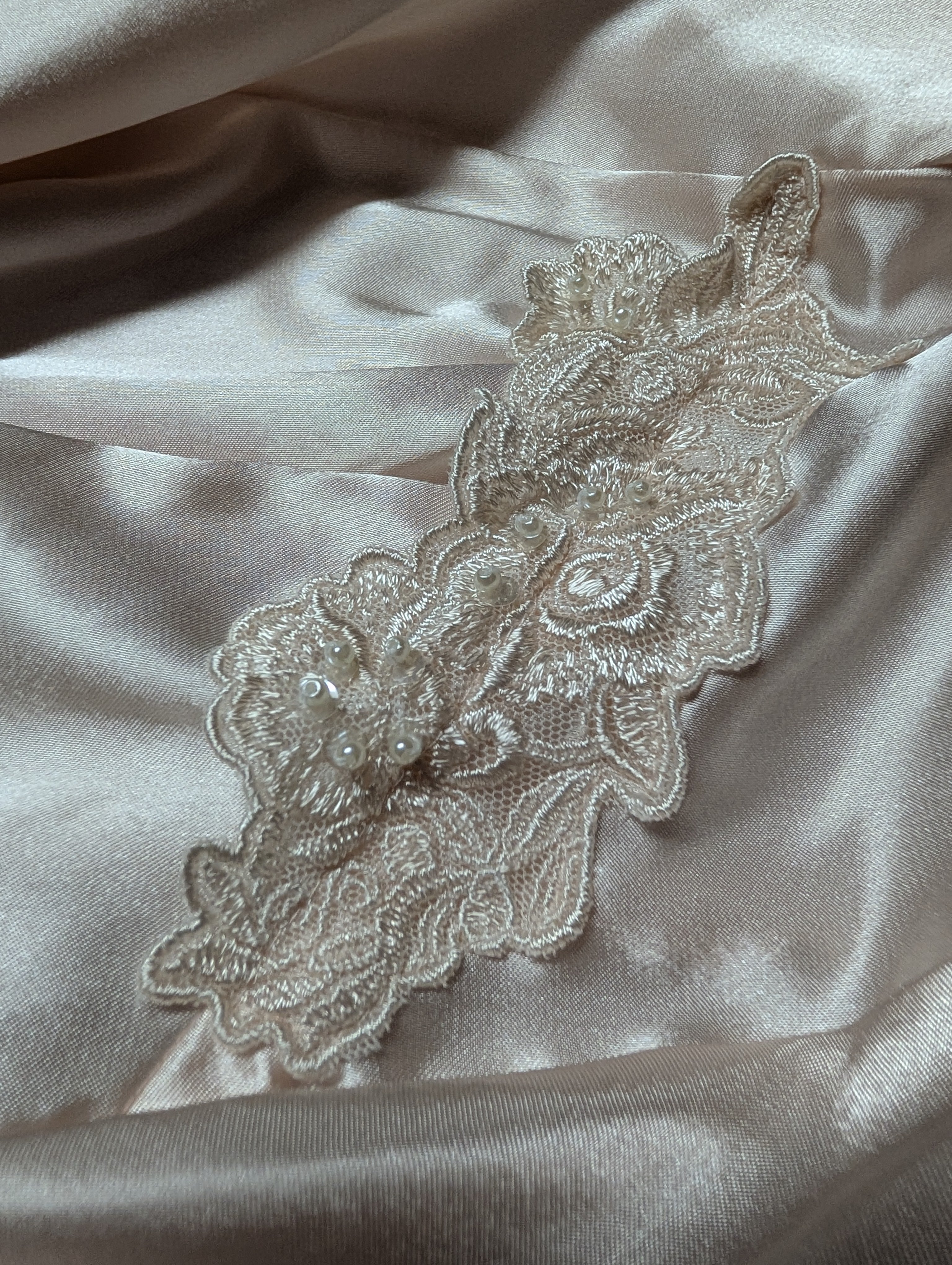 A close-up of a cream-colored floral lace applique with pearl embellishments, laid flat on a draped champagne satin fabric background.