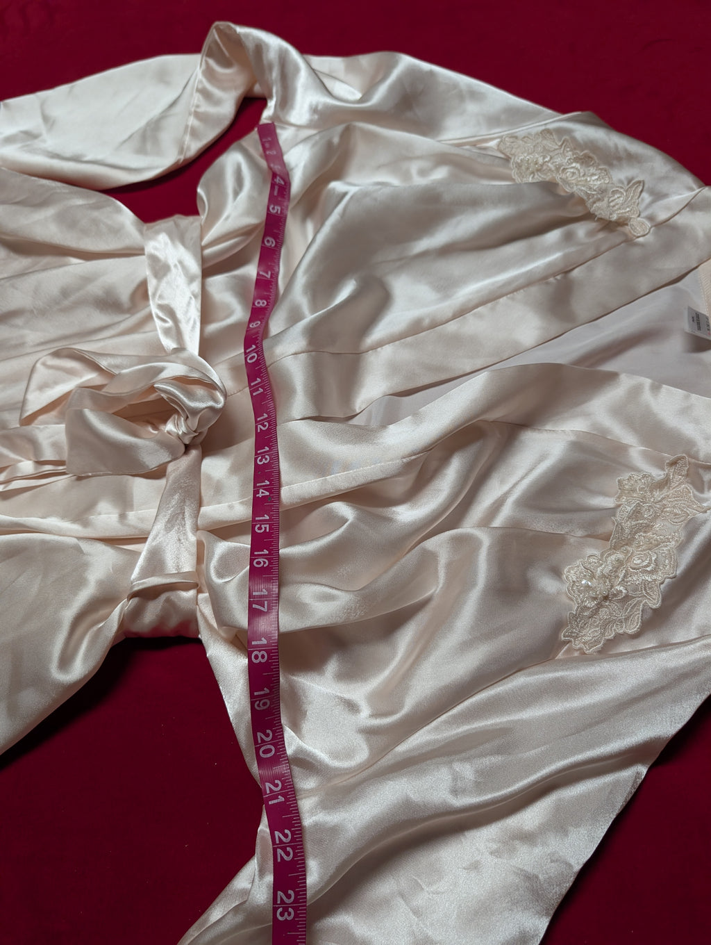 A cream-colored satin robe with lace trim details, laid out on a dark red background with a pink measuring tape draped over it.