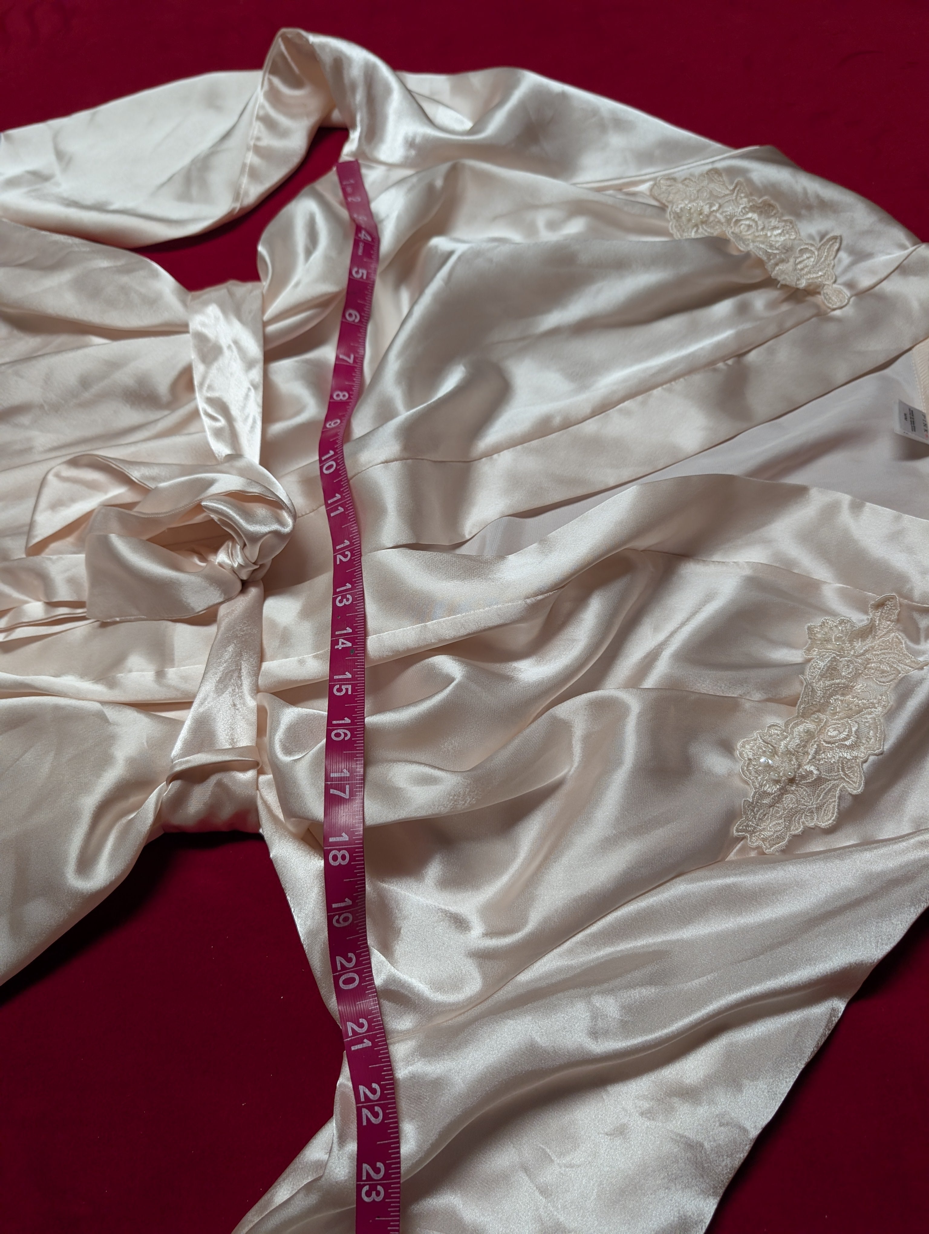 A cream-colored satin robe with lace trim details, laid out on a dark red background with a pink measuring tape draped over it.