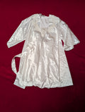 A white satin nightgown with lace trim and a matching tie belt, laid flat on a dark red fabric background.