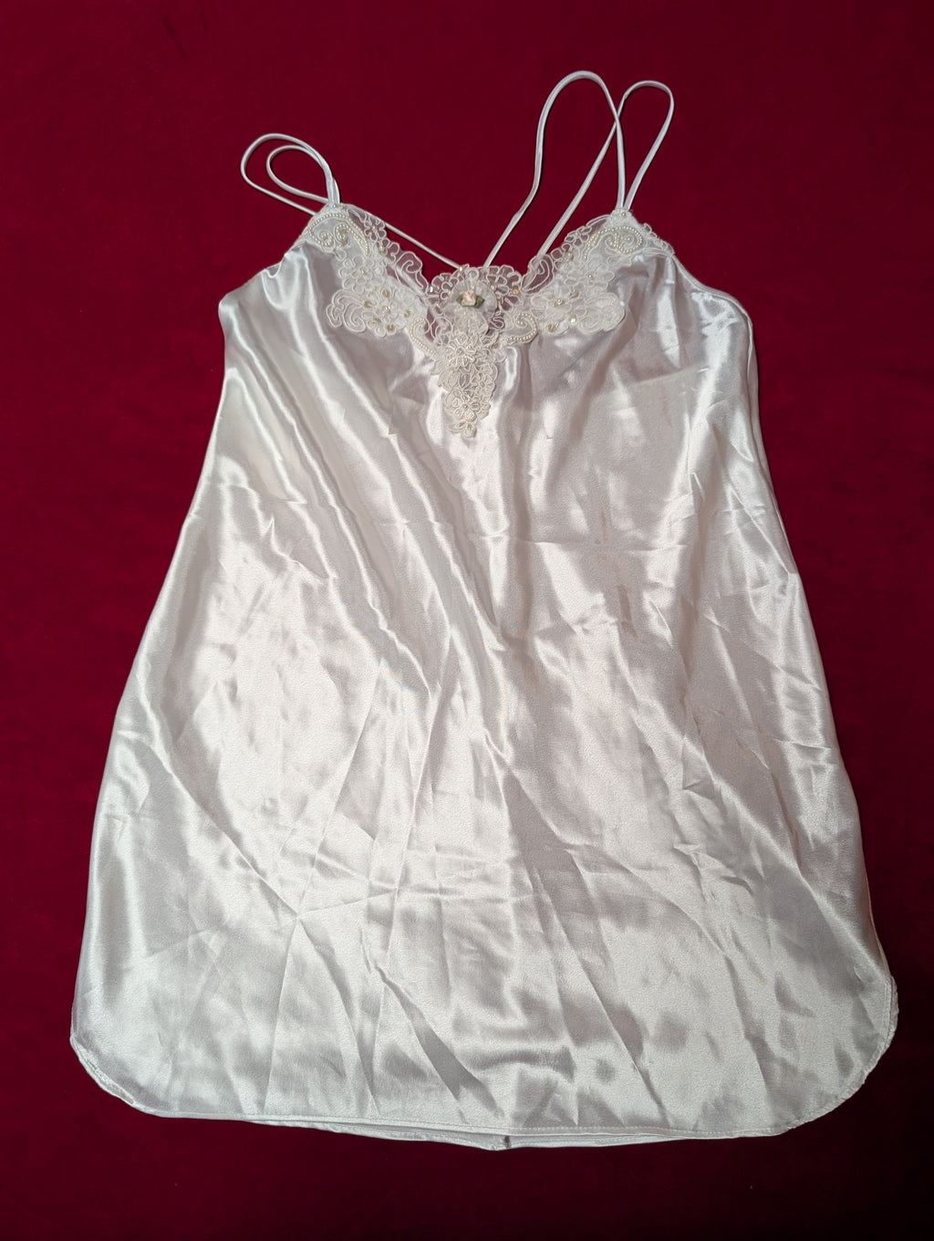 A white satin camisole slip with lace trim and thin adjustable spaghetti straps, laid flat on a dark red background.