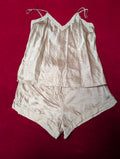 A light beige two-piece lingerie set consisting of a camisole top with thin shoulder straps and matching shorts, laid flat on a dark red background.
