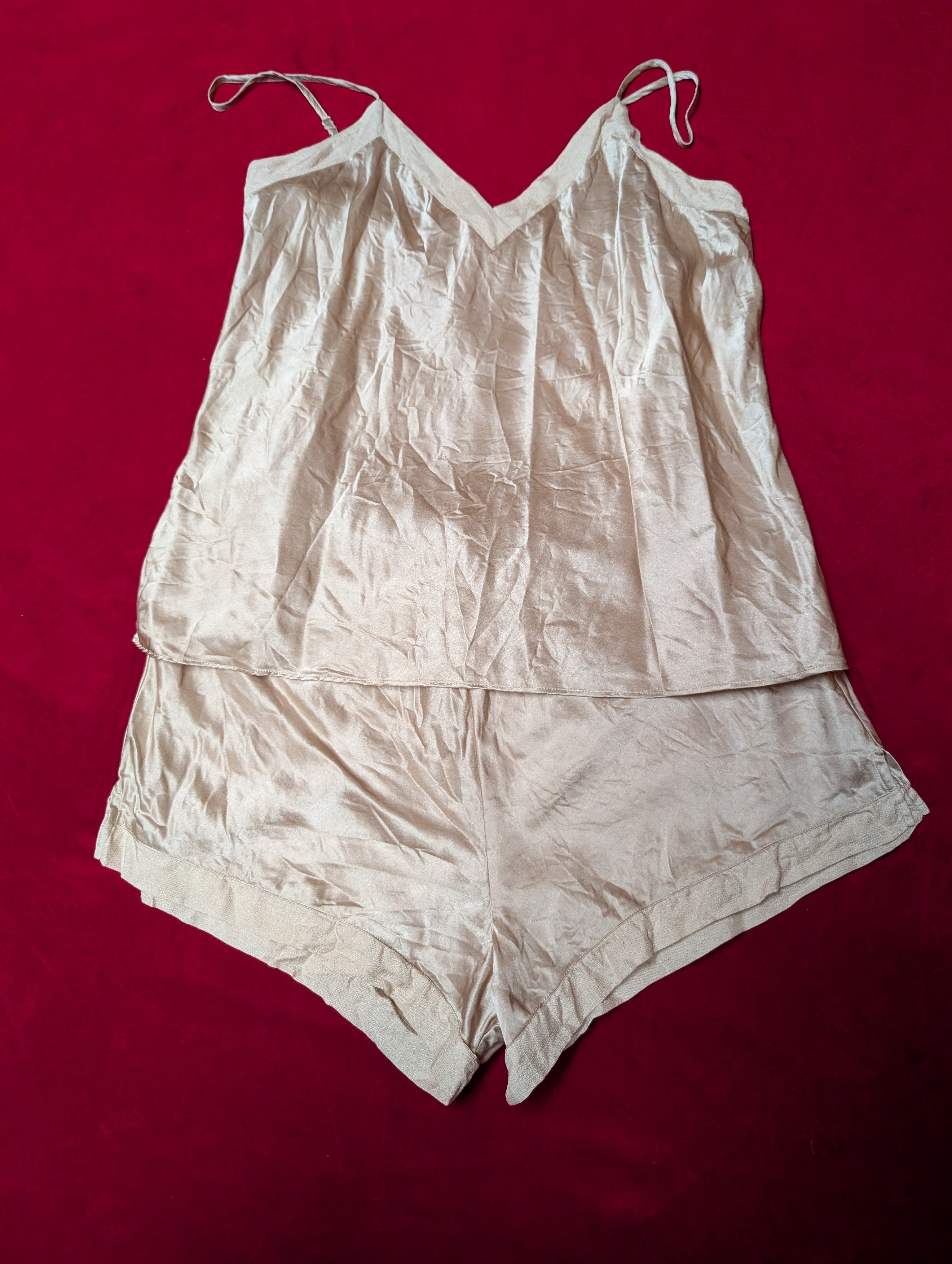 A light beige two-piece lingerie set consisting of a camisole top with thin shoulder straps and matching shorts, laid flat on a dark red background.