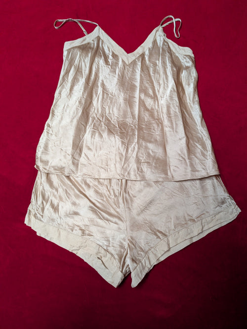 A light beige two-piece lingerie set consisting of a camisole top with thin shoulder straps and matching shorts, laid flat on a dark red background.