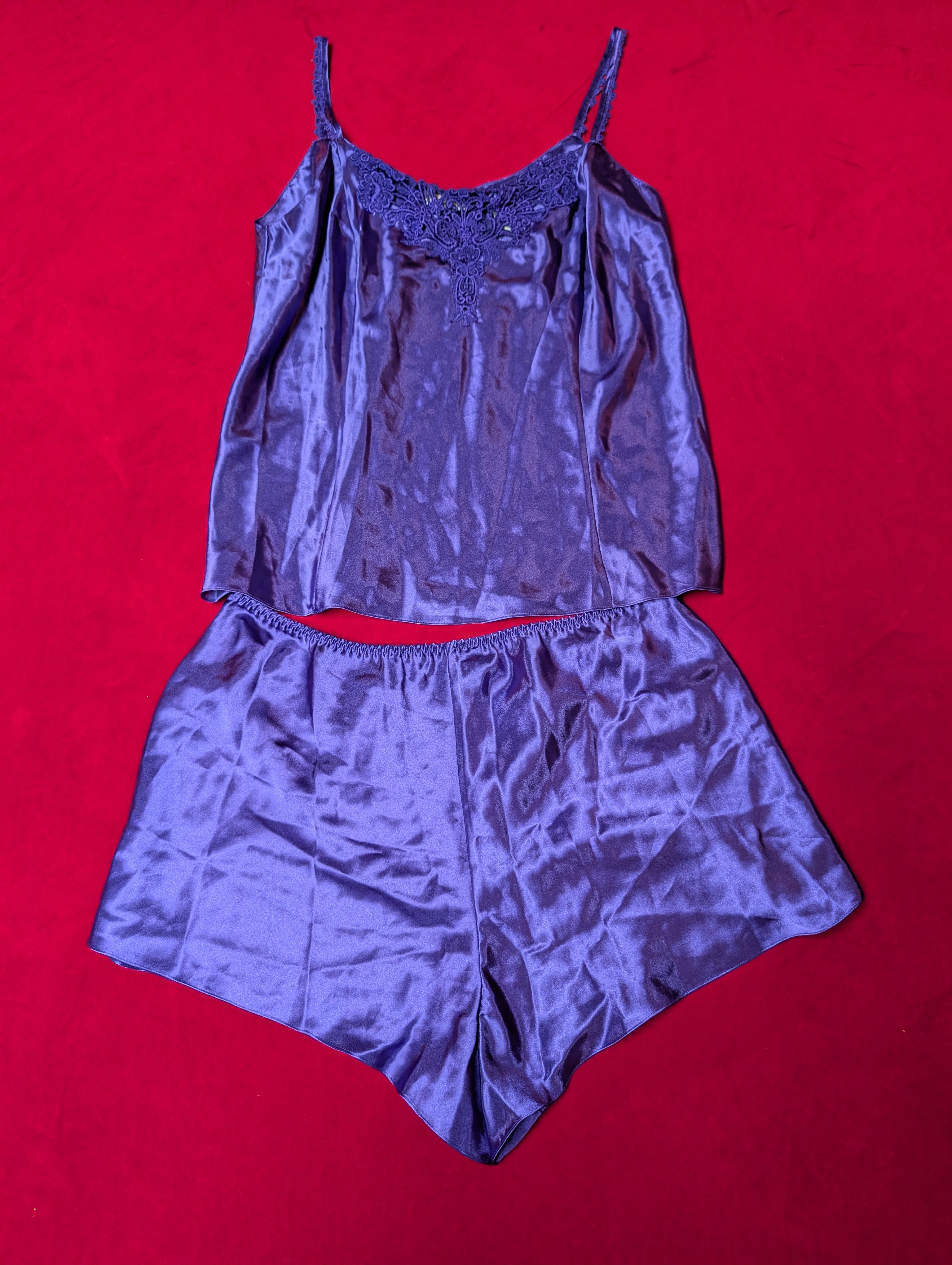 A two-piece satin pajama set in a shimmering purple-blue color, featuring a camisole top with thin straps and a matching short set, laid flat on a bright red background.