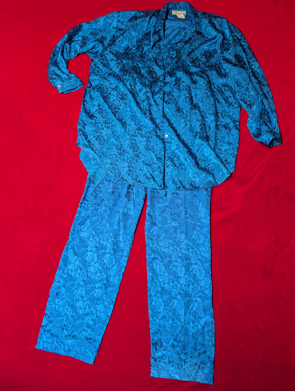 A two-piece pajama set consisting of a long-sleeve button-up shirt and matching pants, both in a vibrant blue and black floral jacquard pattern, laid flat on a bright red background.
