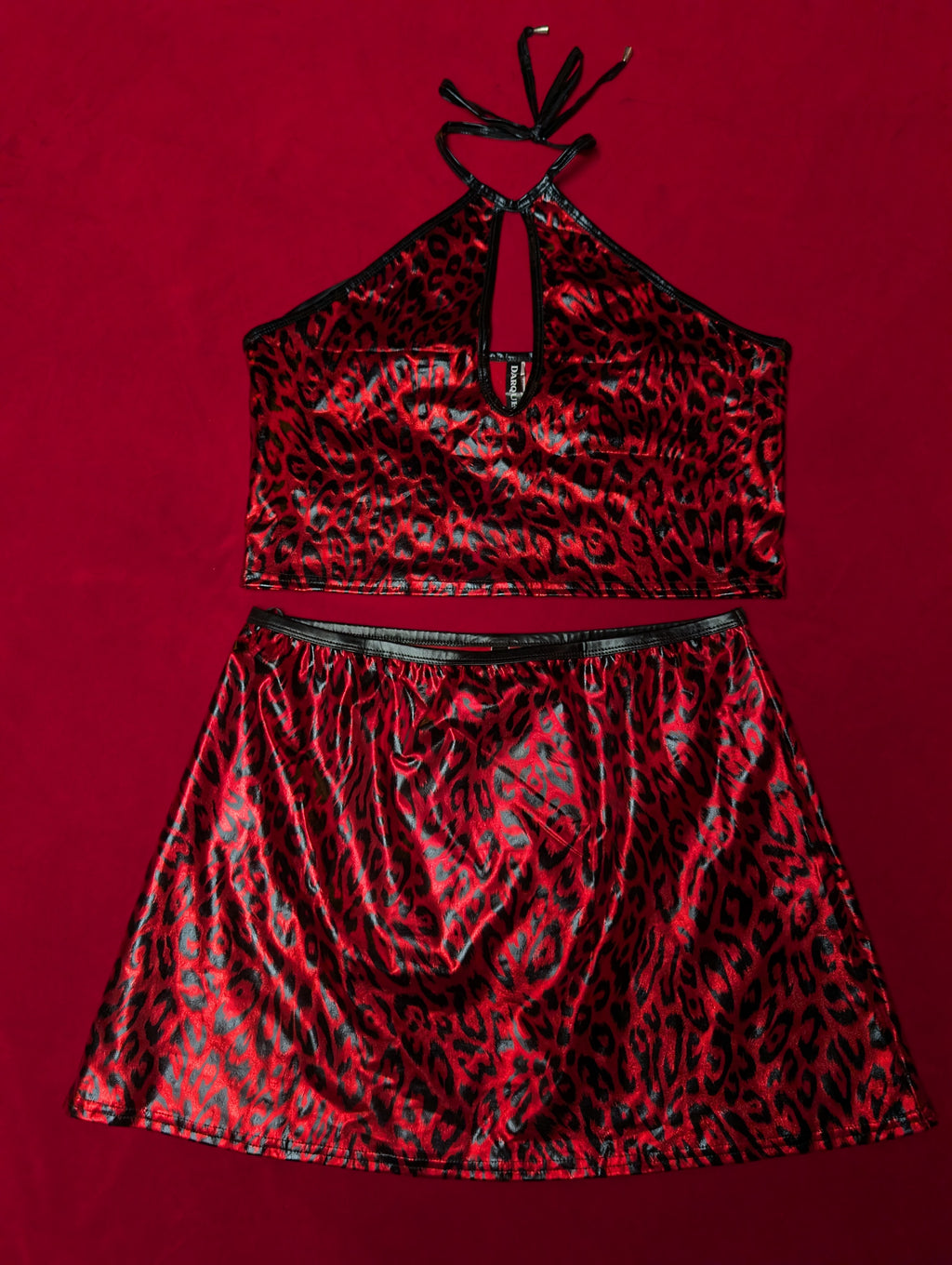 A two-piece halter crop top and matching mini skirt set in a shiny red and black leopard print pattern, displayed flat against a solid red background.