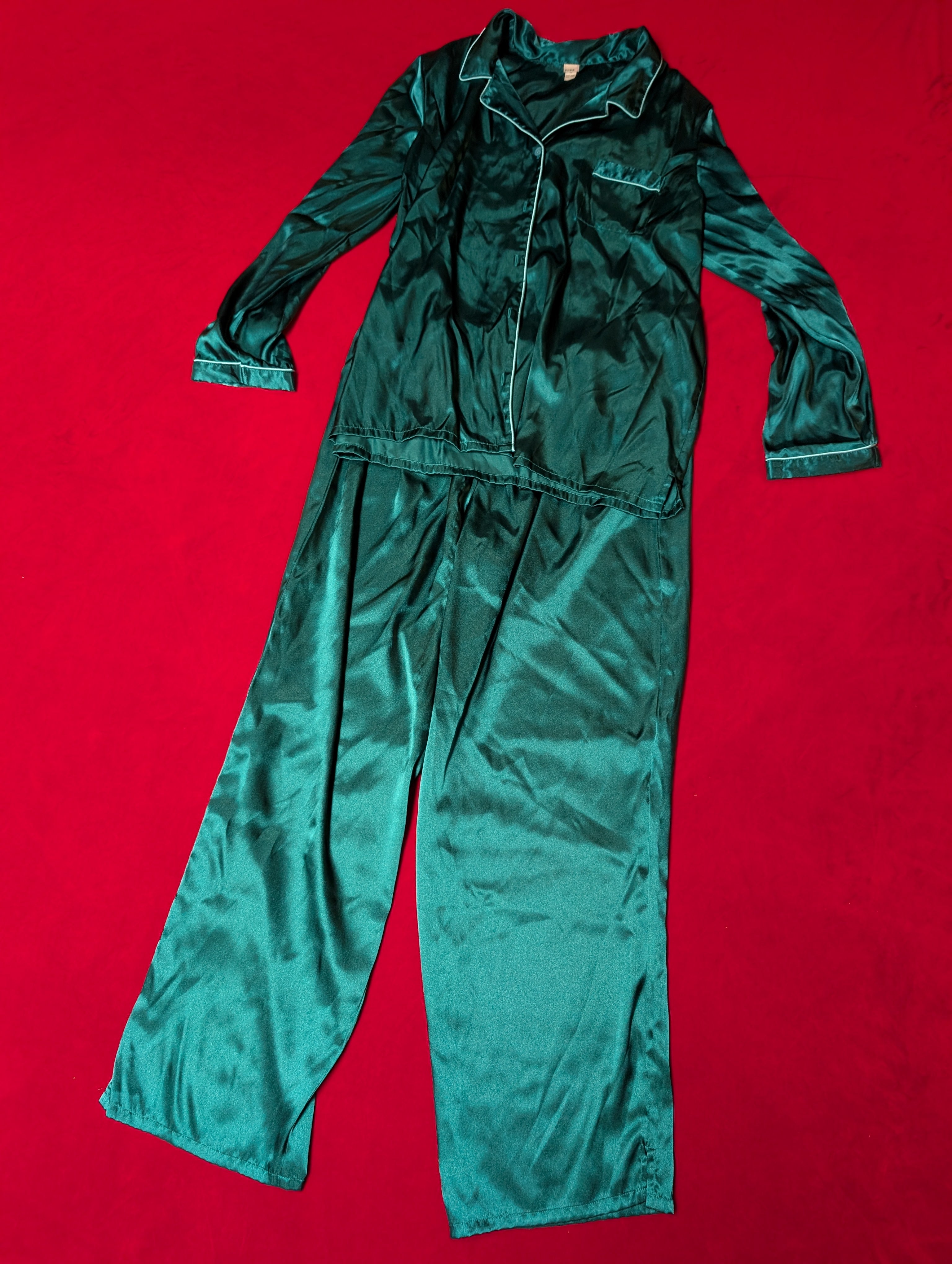 A two-piece satin pajama set in a deep teal green color, laid flat on a bright red background. The set includes a long-sleeve button-up top with a collar and matching pants.