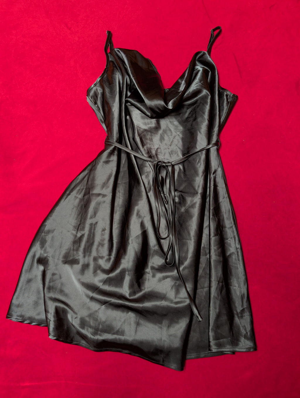 A black satin slip dress with spaghetti straps and a tie waist, laid flat on a vibrant red background.