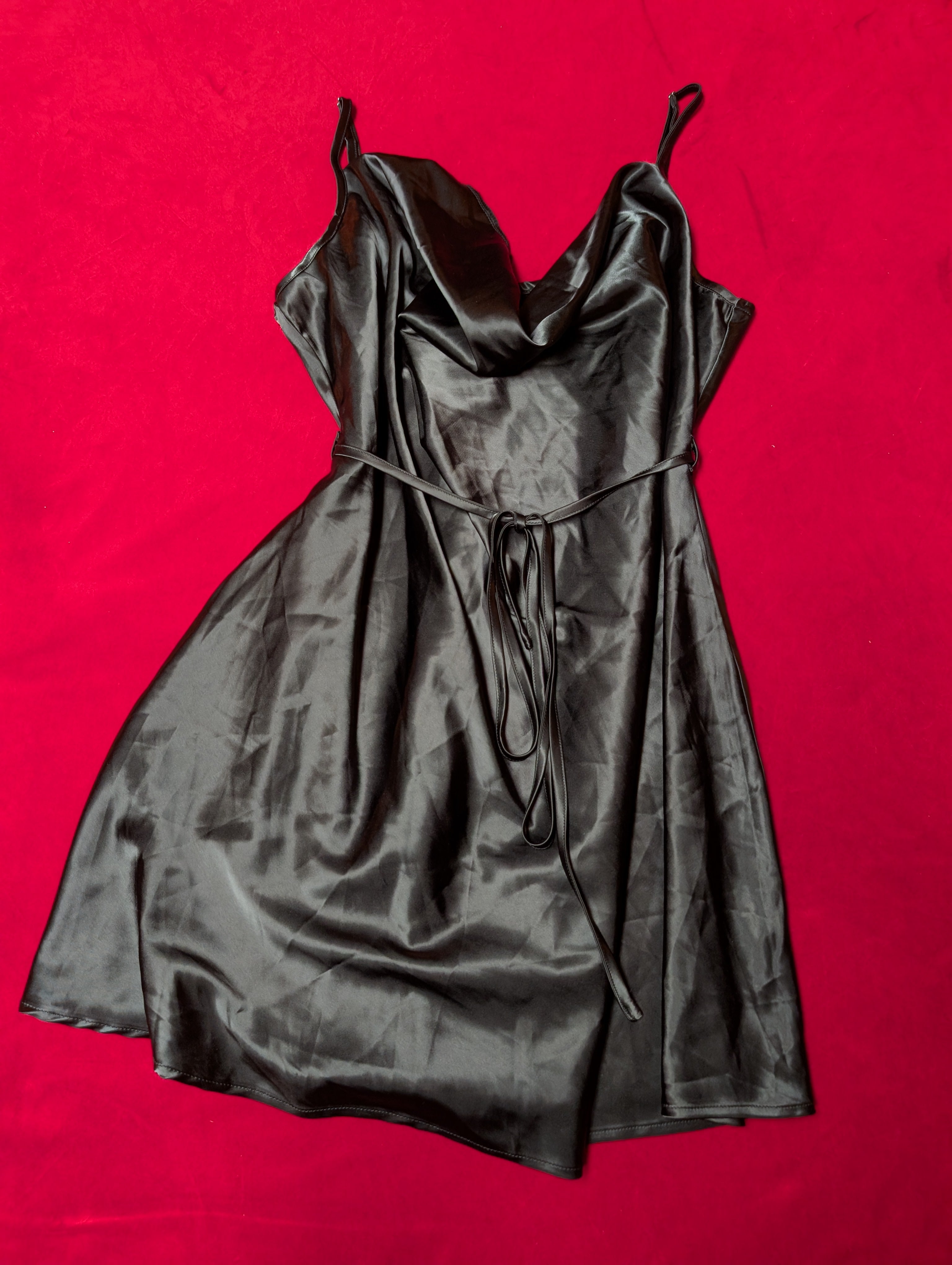 A black satin slip dress with spaghetti straps and a tie waist, laid flat on a vibrant red background.