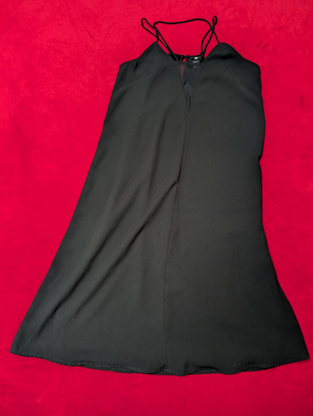 A black sleeveless slip dress with thin shoulder straps and a V-neckline, laid flat on a vibrant red textured background.