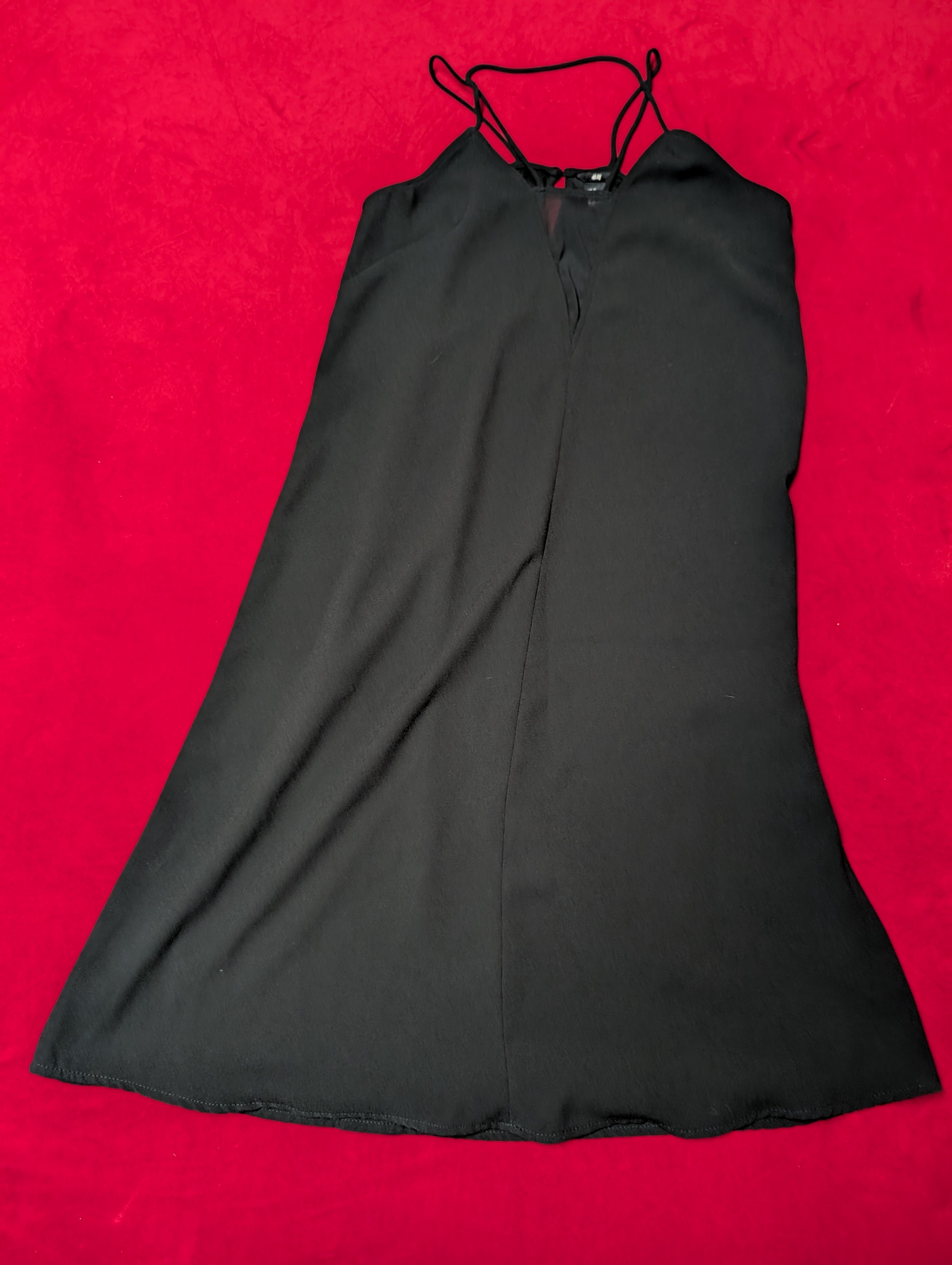 A black sleeveless slip dress with thin shoulder straps and a V-neckline, laid flat on a vibrant red textured background.