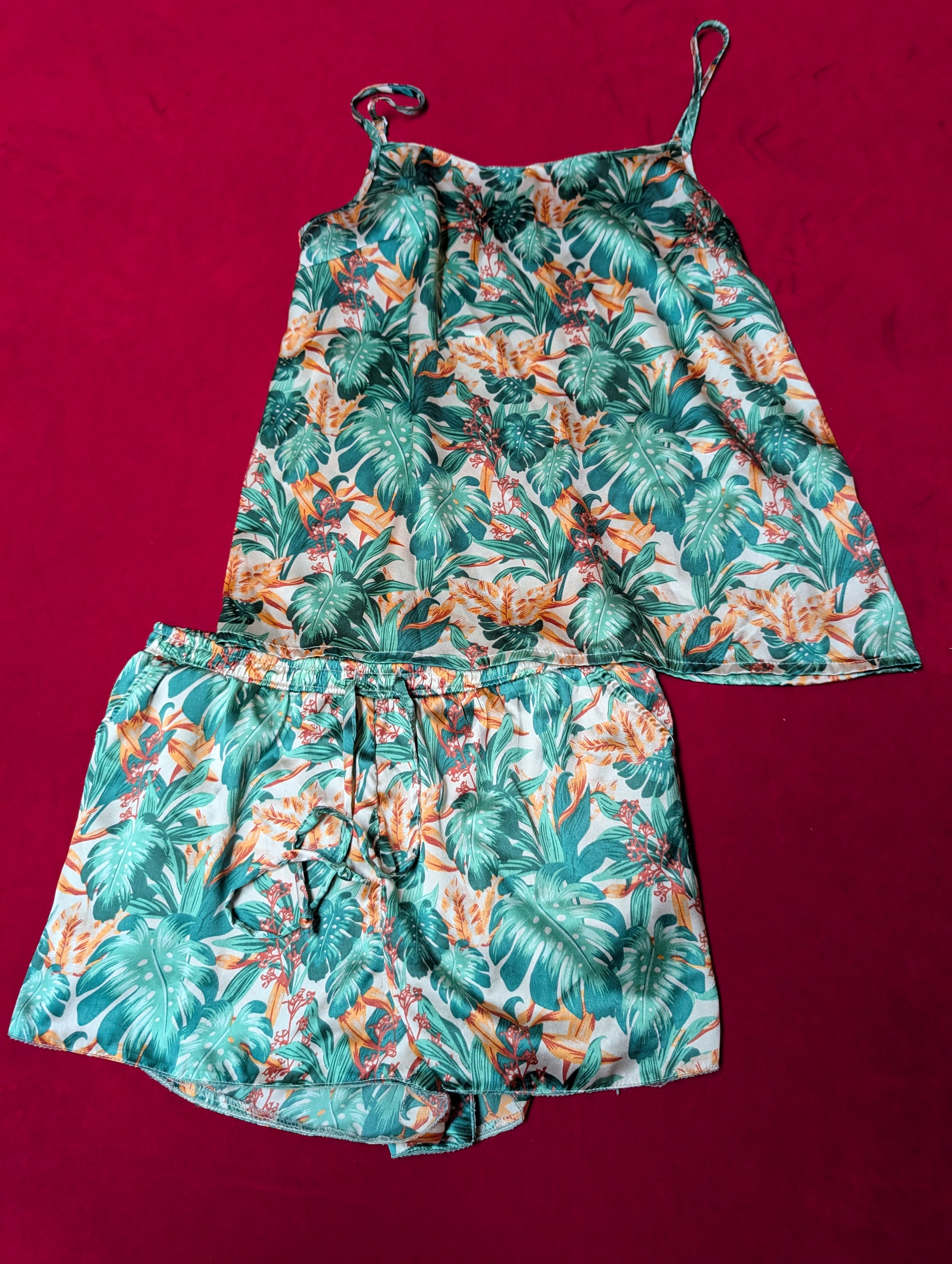 A two-piece tropical print set consisting of a sleeveless camisole top and matching shorts, laid flat on a dark red background. The print features green palm leaves and orange flowers on a white base.