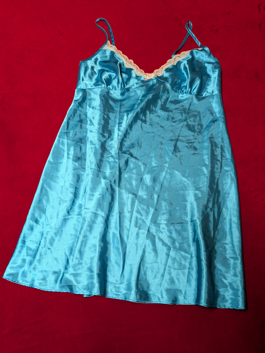 A light blue satin slip dress with thin shoulder straps and a peach-colored lace trim along the neckline, laid flat on a red background.