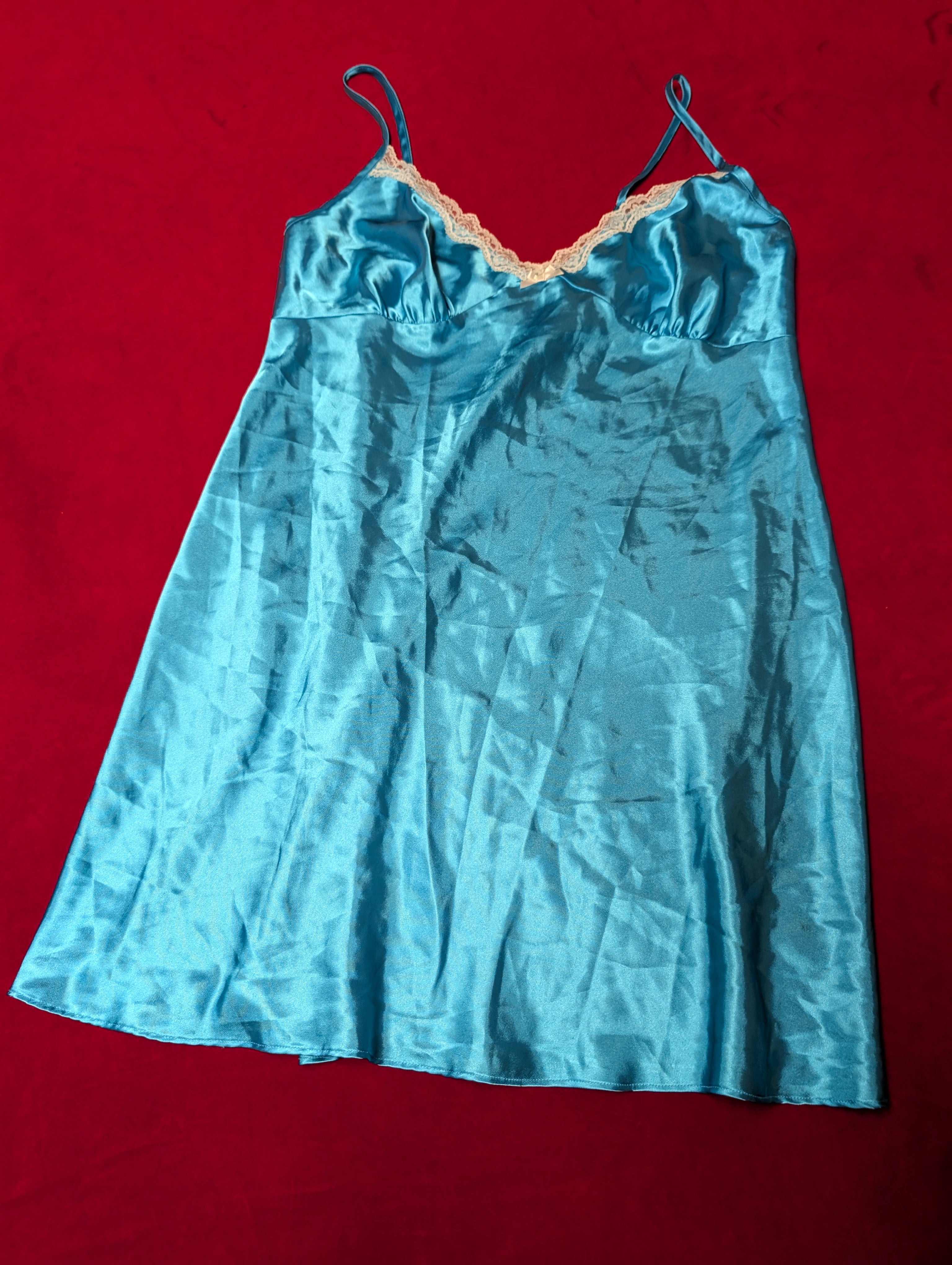 A light blue satin slip dress with thin shoulder straps and a peach-colored lace trim along the neckline, laid flat on a red background.