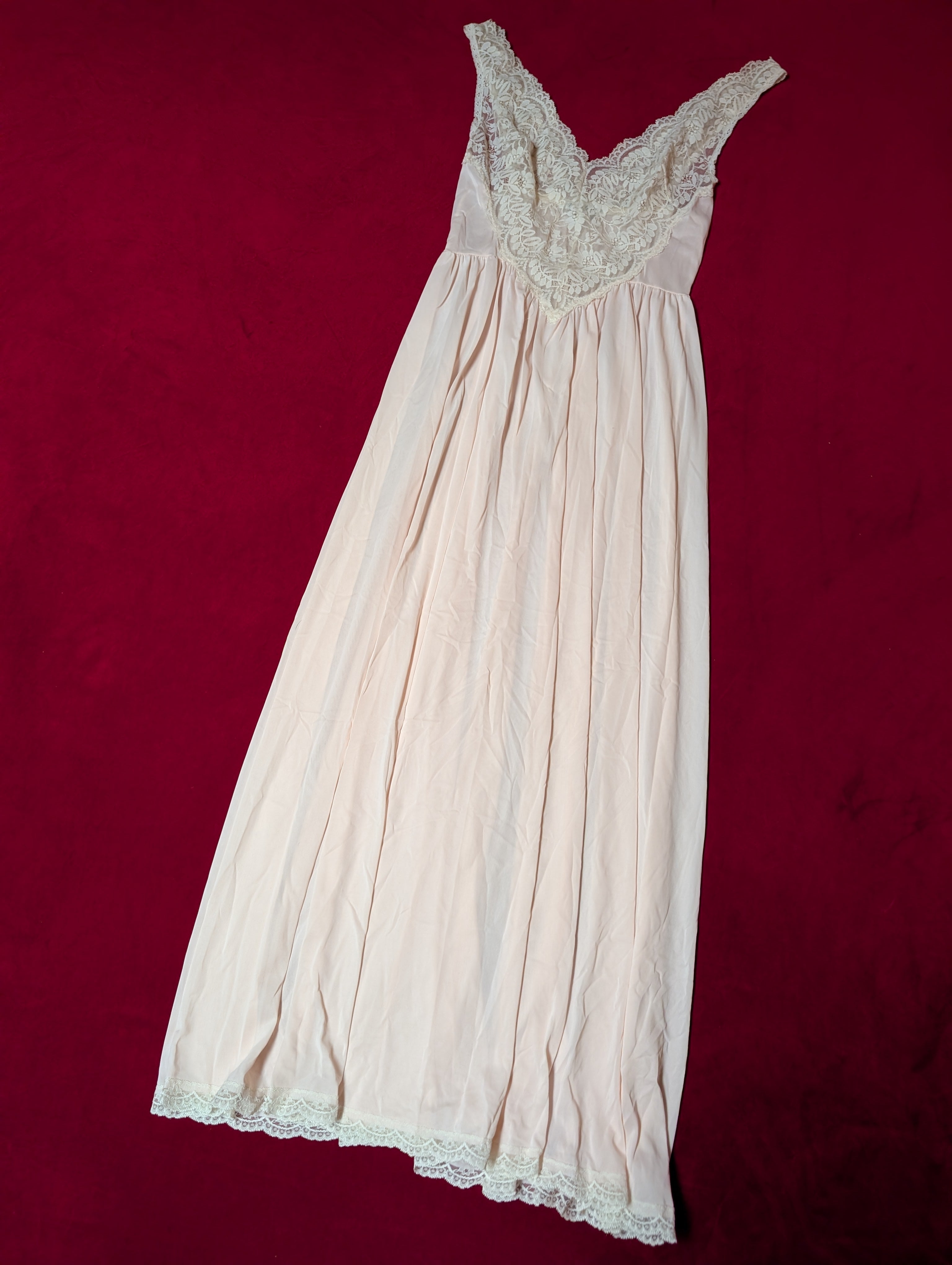 A pale pink or cream-colored nightgown with a lace-trimmed scoop neckline, laid flat on a dark red background.
