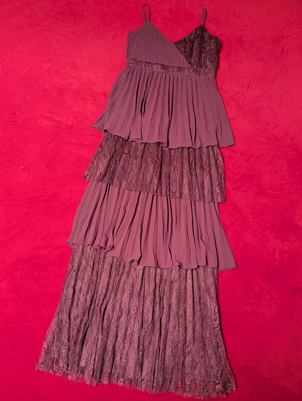 A tiered pleated dress in a deep red or plum purple color, laid flat on a vibrant hot red textured background.