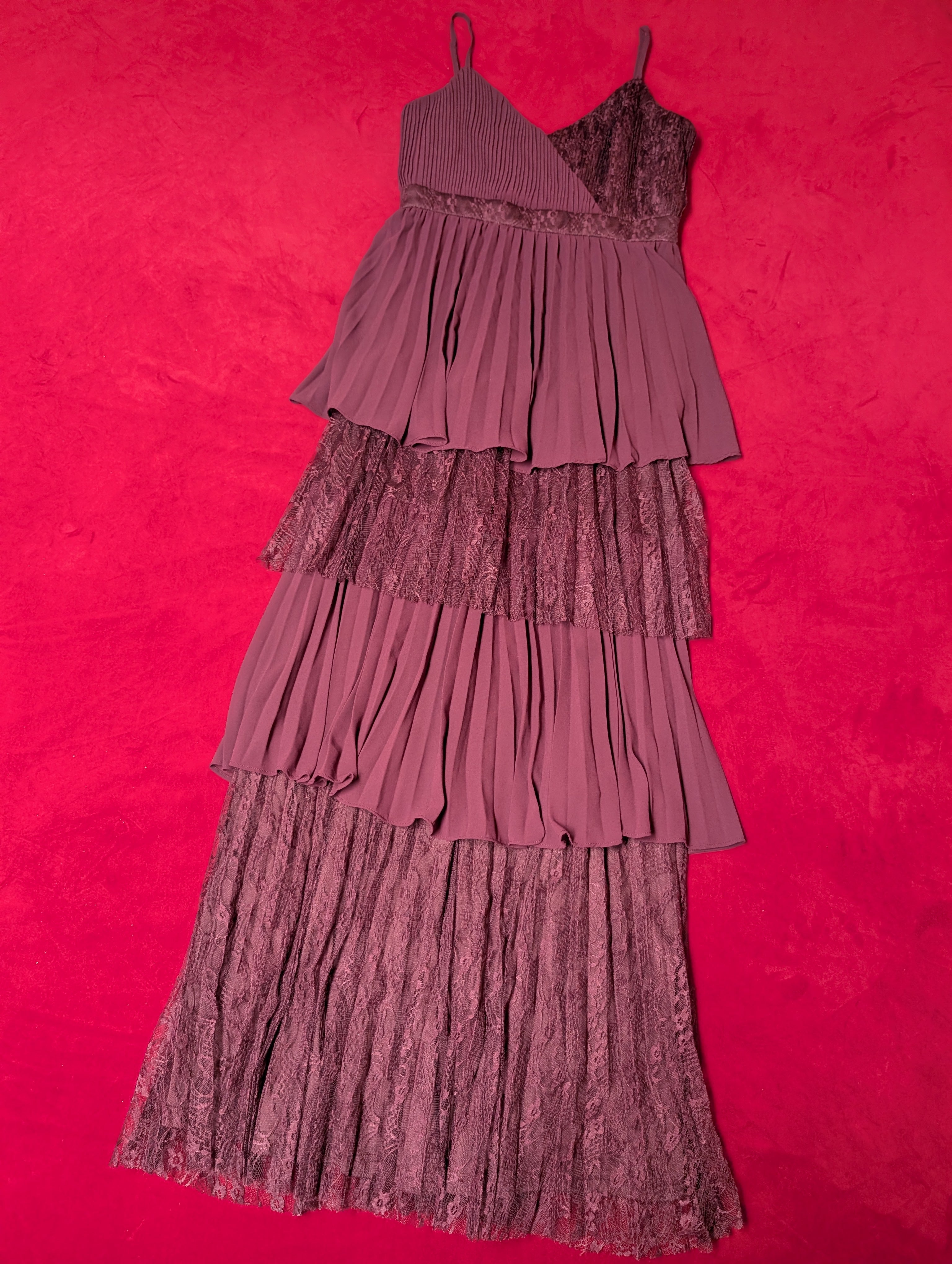 A tiered pleated dress in a deep red or plum purple color, laid flat on a vibrant hot red textured background.