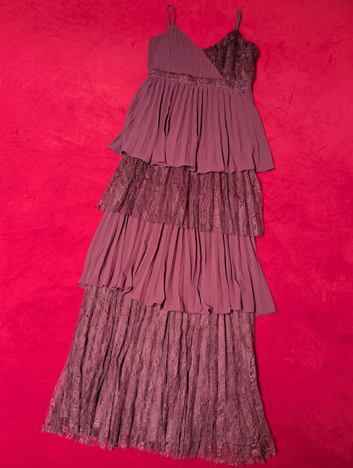A tiered pleated dress in a deep red or plum purple color, laid flat on a vibrant hot red textured background.