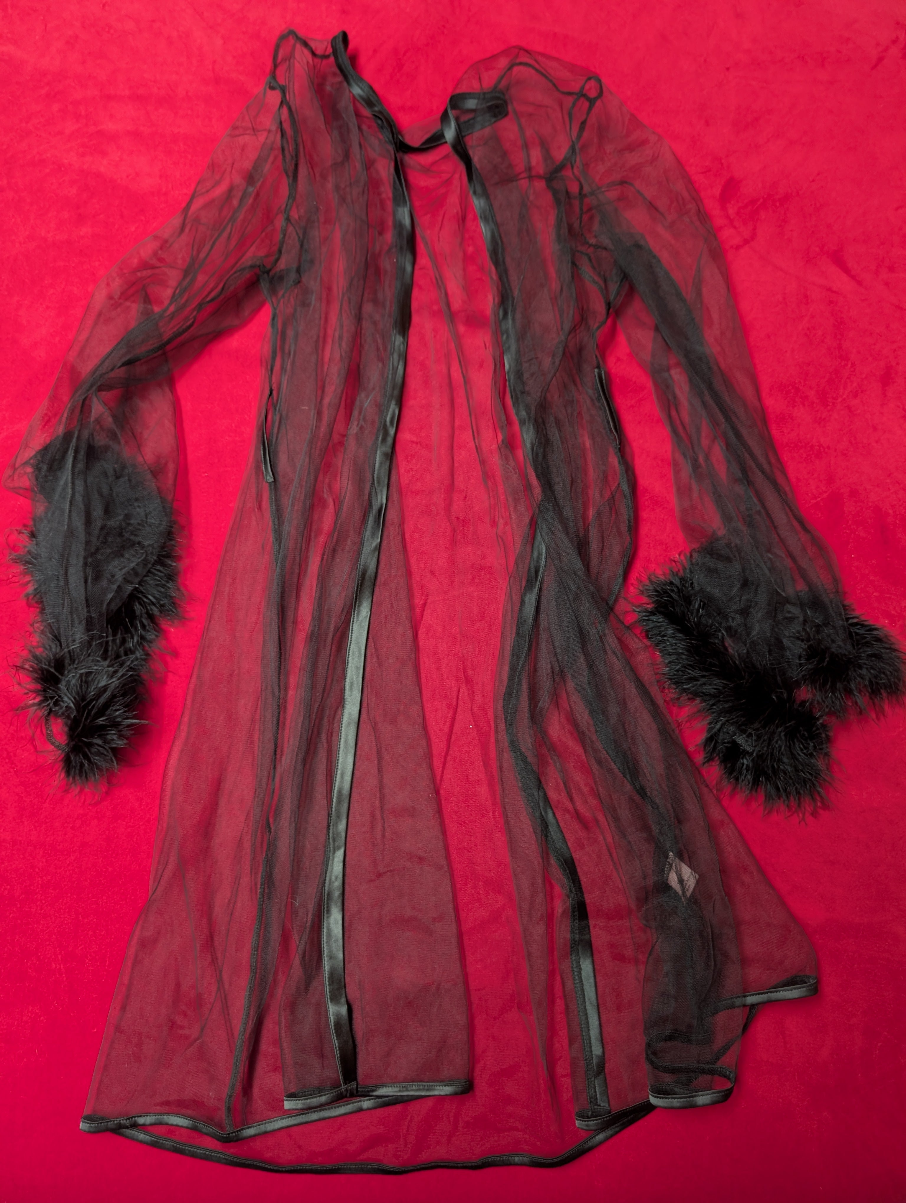 A black sheer tulle robe or long coat with black feather trim at the cuffs, laid flat on a bright red background.