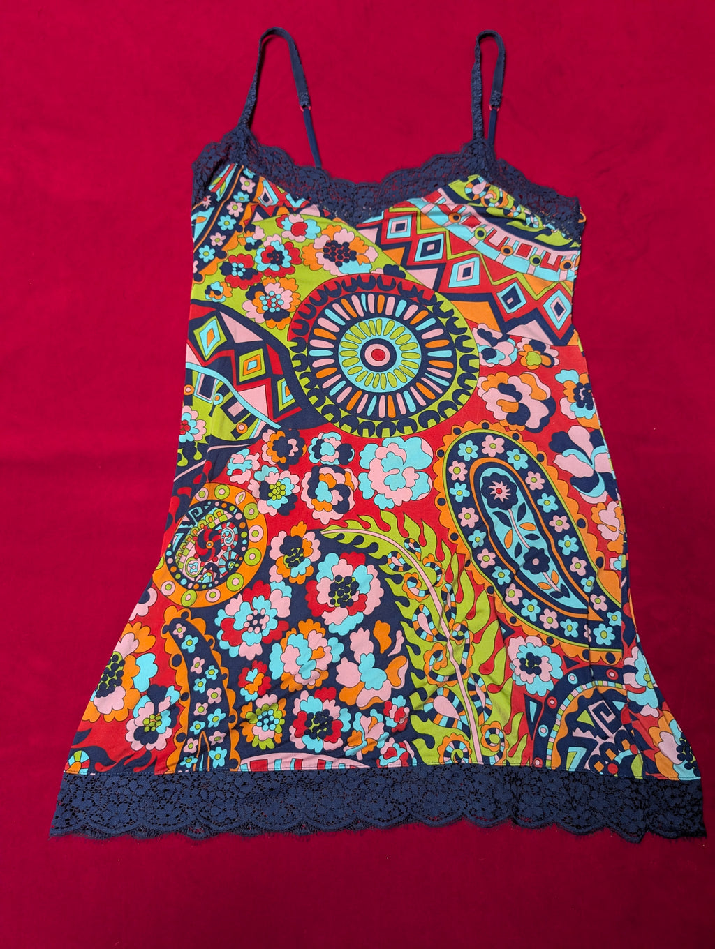 A colorful spaghetti-strap camisole dress featuring a vibrant bohemian paisley and floral print, finished with dark navy lace trim at the neckline and hem, laid flat on a red background.