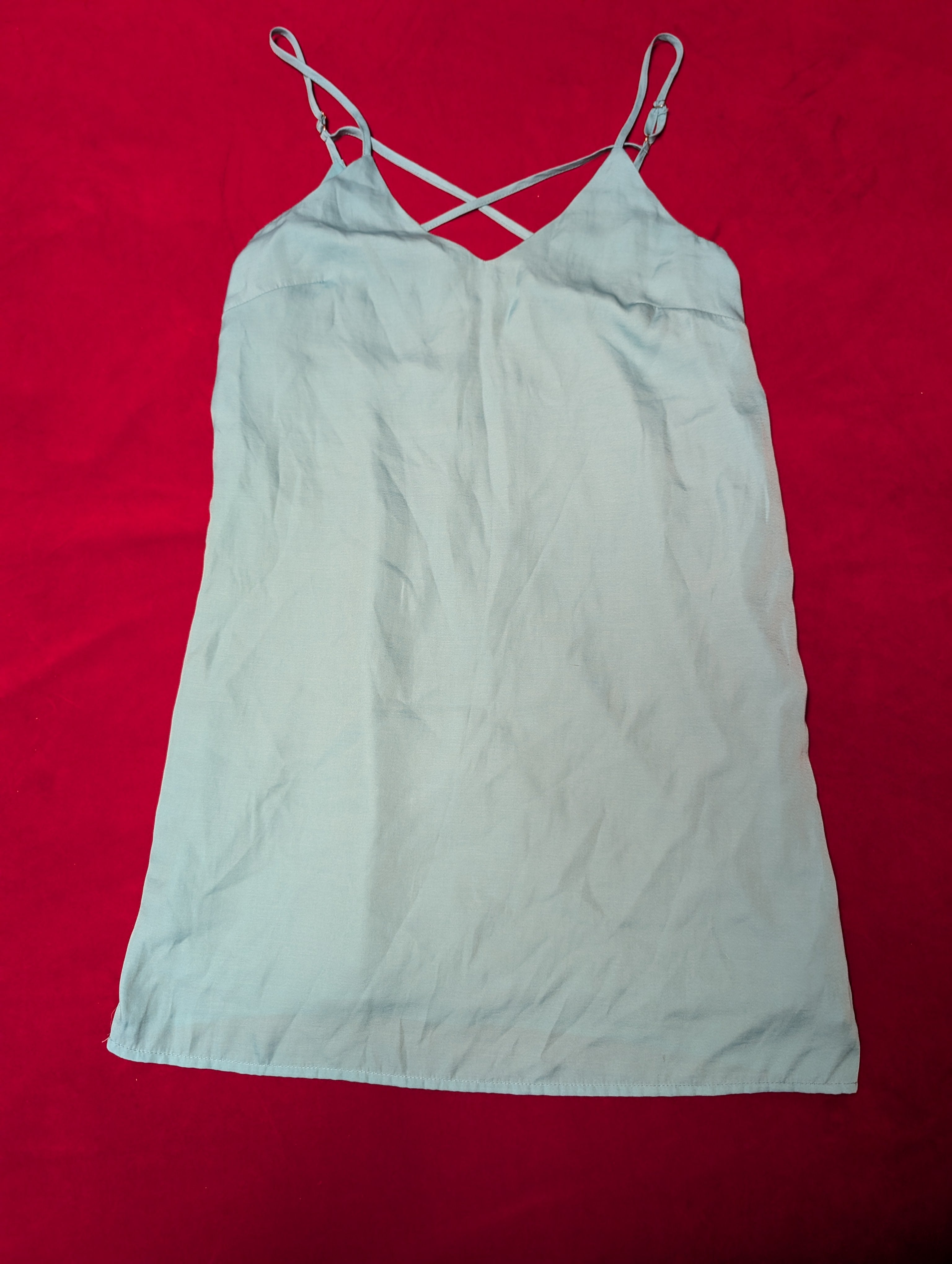 A light sky blue or pale gray-blue sleeveless camisole dress with thin adjustable spaghetti straps and a crisscross back design, laid flat on a vibrant red background.
