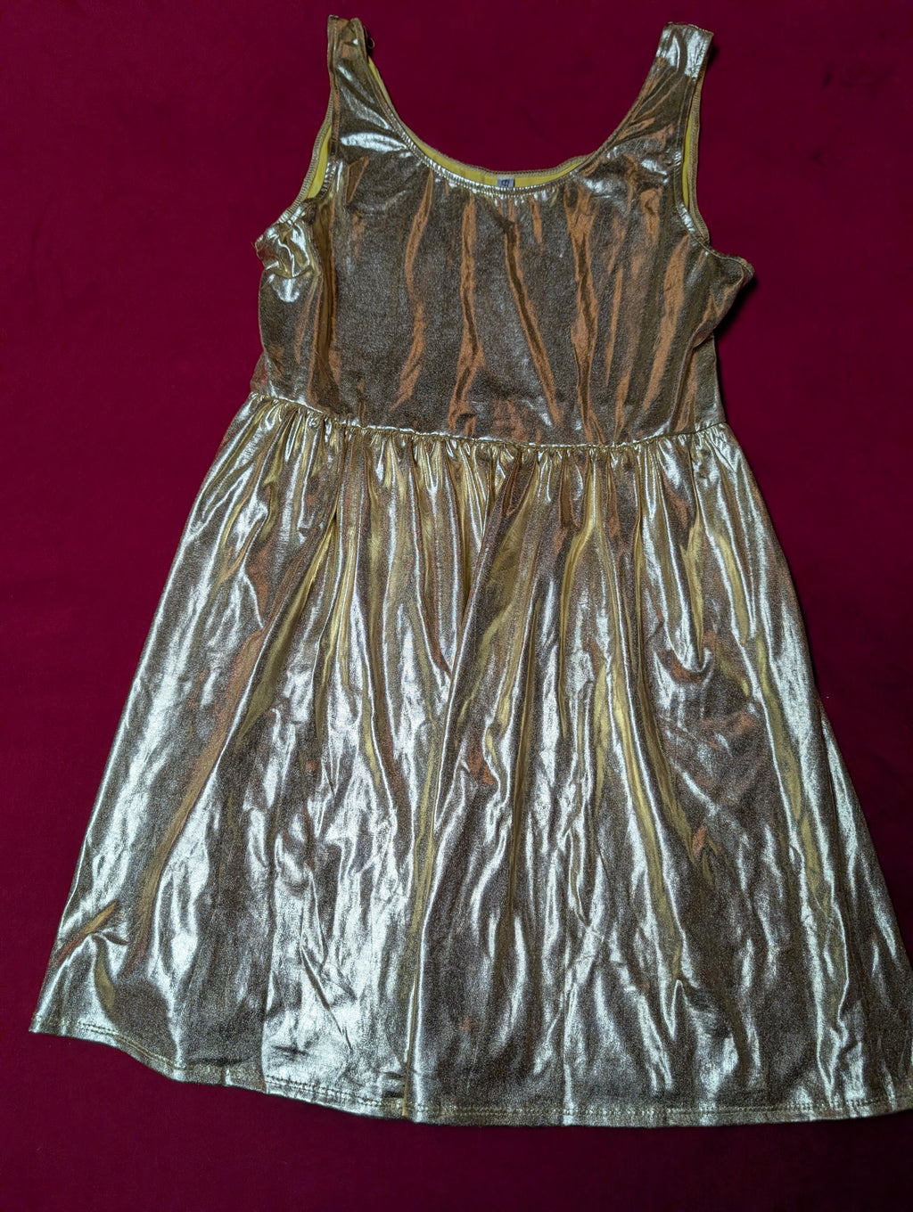 A shiny metallic gold sleeveless dress with a gathered waist, laid flat on a dark red background.