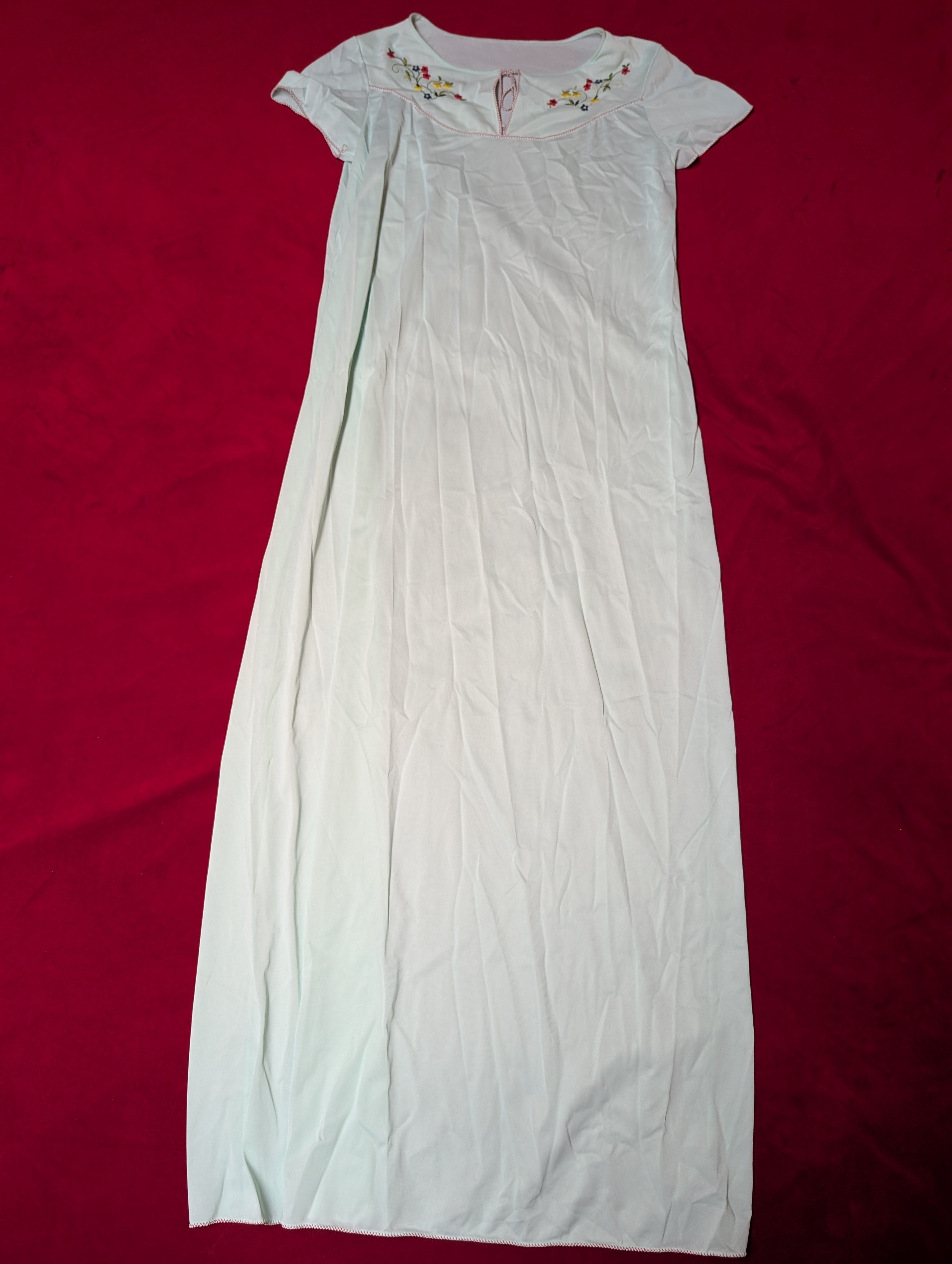 A mint green short-sleeve nightgown with delicate multicolor folk floral embroidery on the chest, laid flat on a vibrant red background.