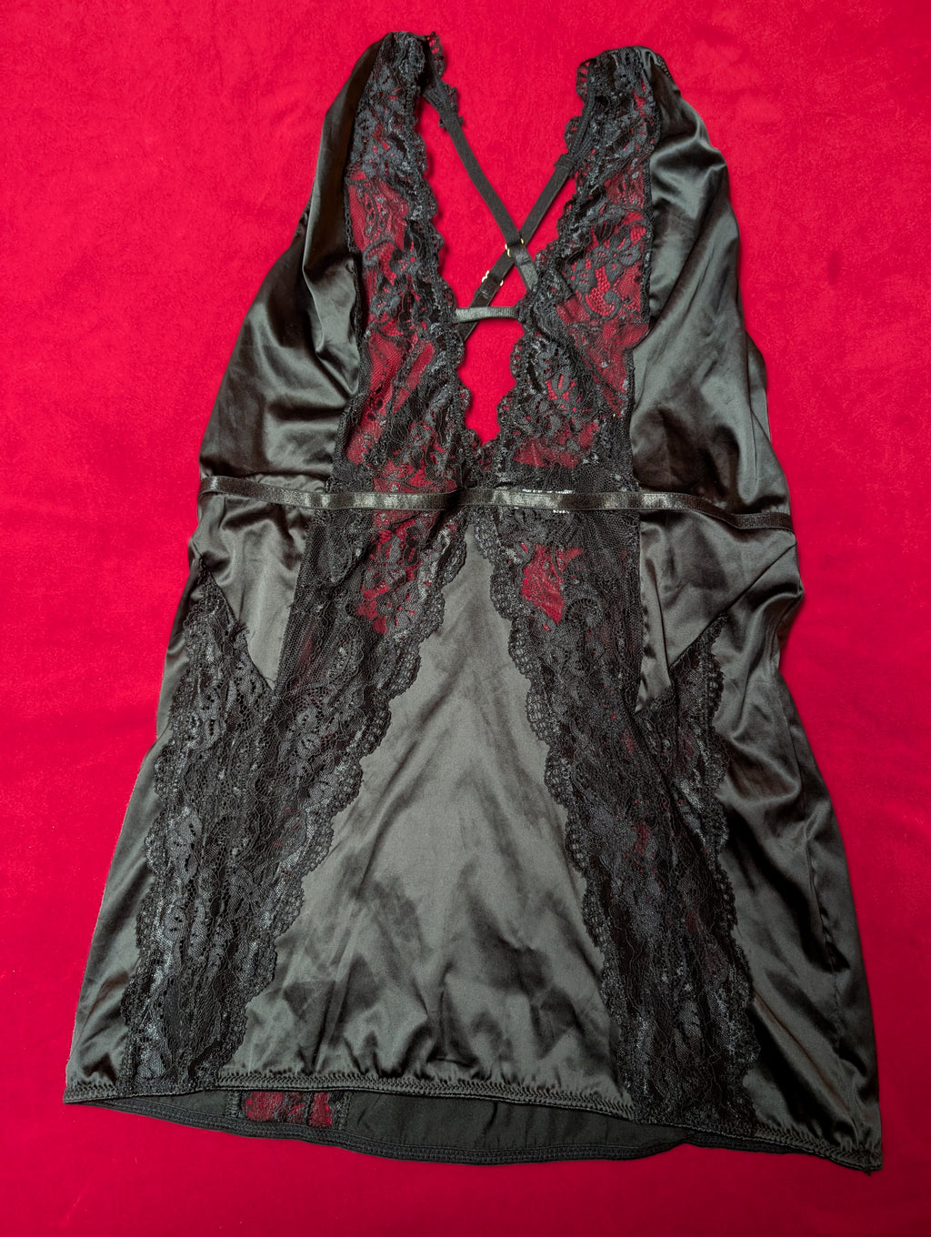 A black satin chemise with black lace panels and a criss-cross lace back, laid flat on a bright red background.