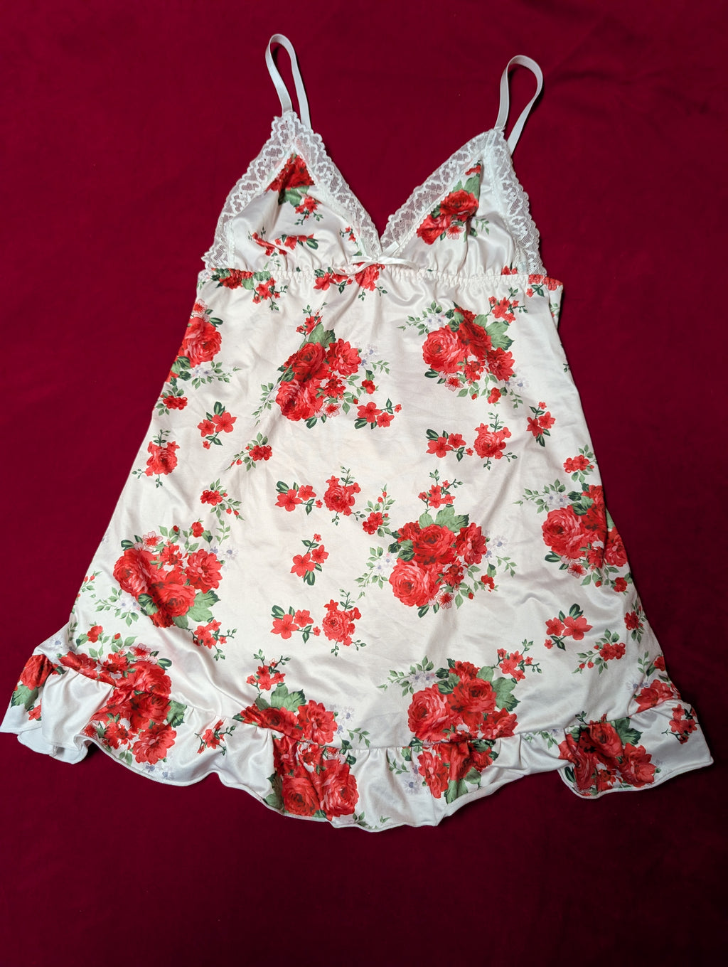 A white camisole nightgown with a red rose floral print and lace trim, laid flat on a dark red background.
