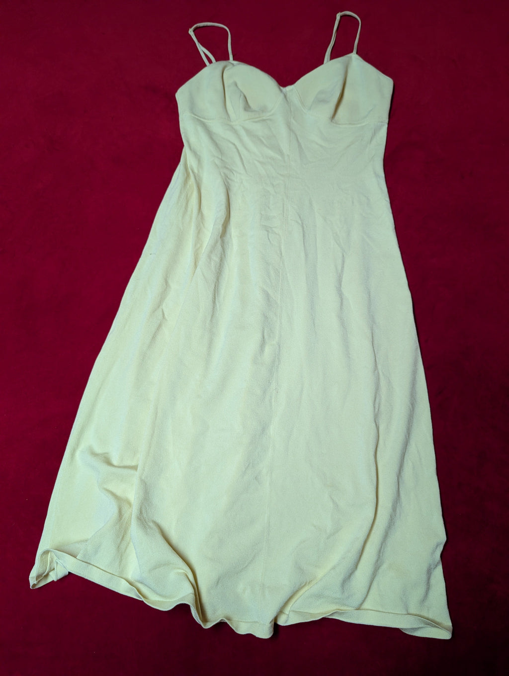 A butter-yellow colored sleeveless slip dress or long camisole with thin spaghetti straps, laid flat on a dark red background.