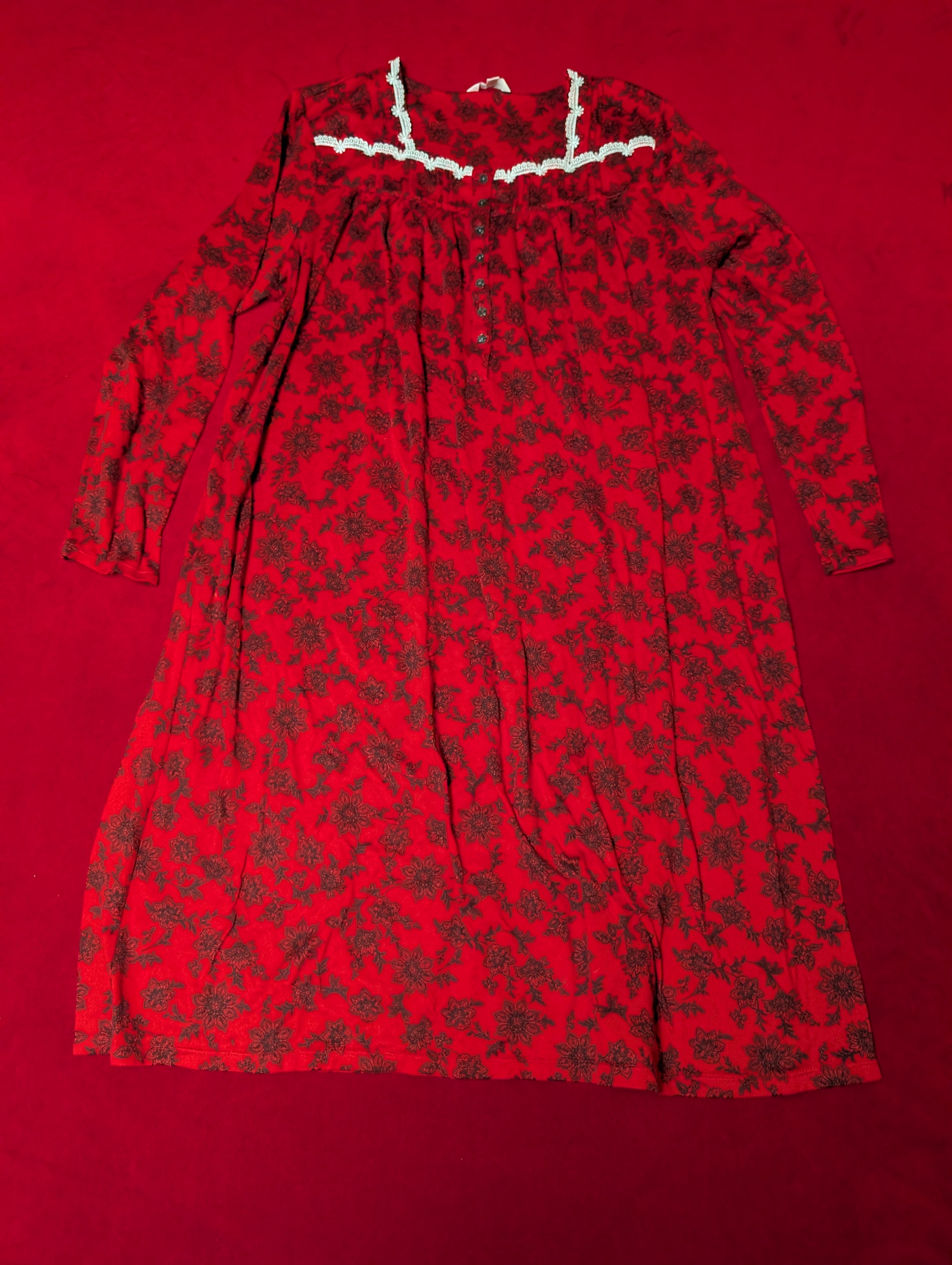 A red long-sleeve night dress with a dark floral pattern, laid flat on a solid red background. The dress features a white lace collar trim and buttons.