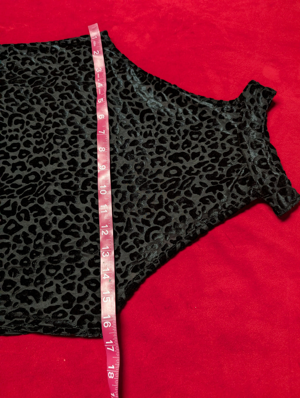 A black leopard print garment laid flat on a bright red background, with a pink measuring tape running vertically through the center to show dimensions.