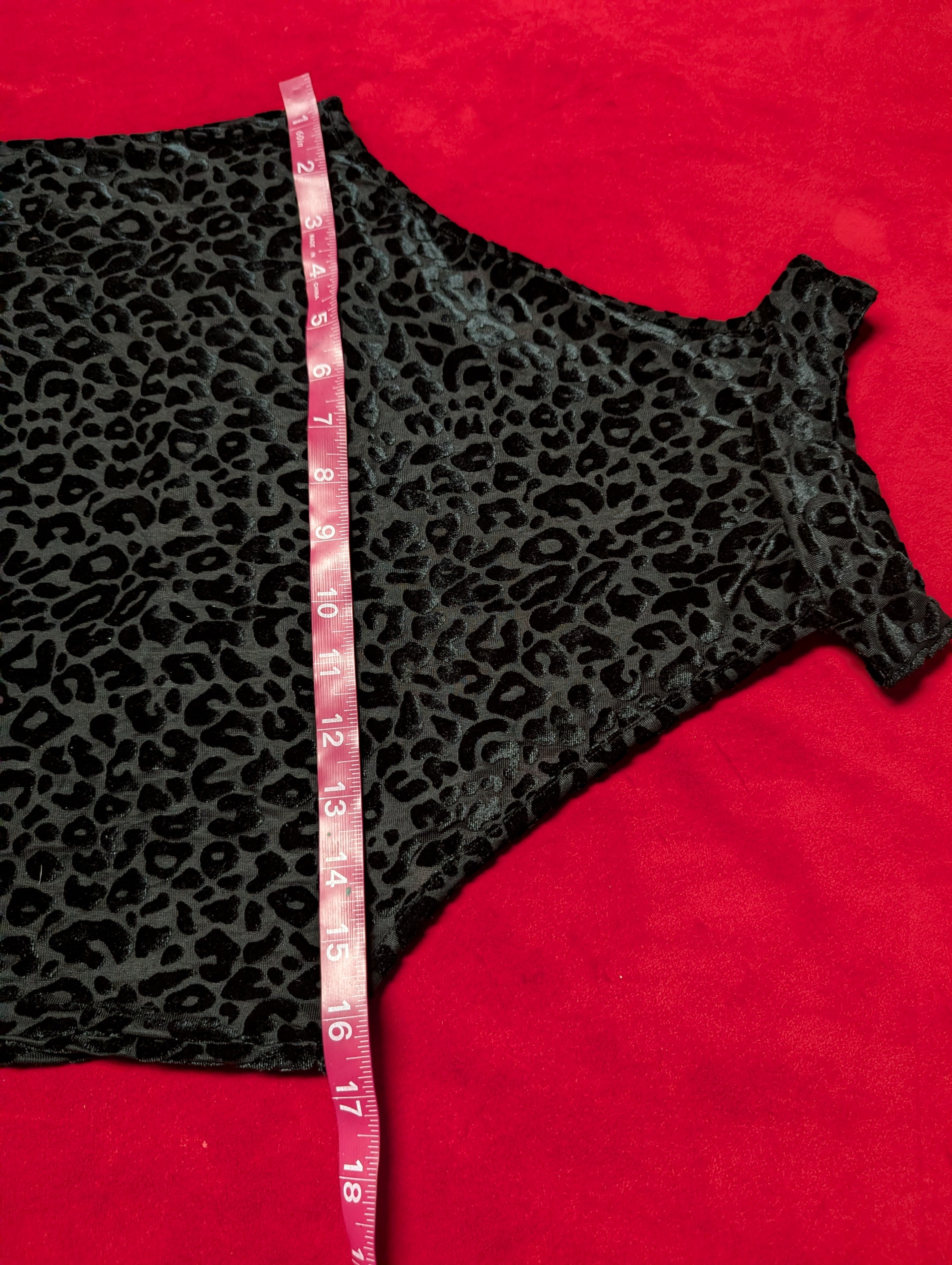 A black leopard print garment laid flat on a bright red background, with a pink measuring tape running vertically through the center to show dimensions.
