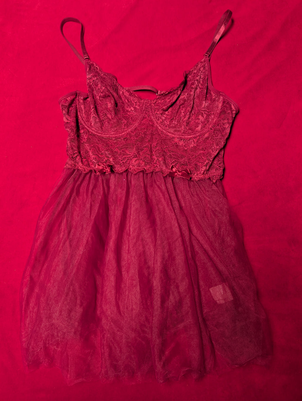 A dark red lace and tulle slip dress with thin spaghetti straps, laid flat on a vibrant red fabric background.