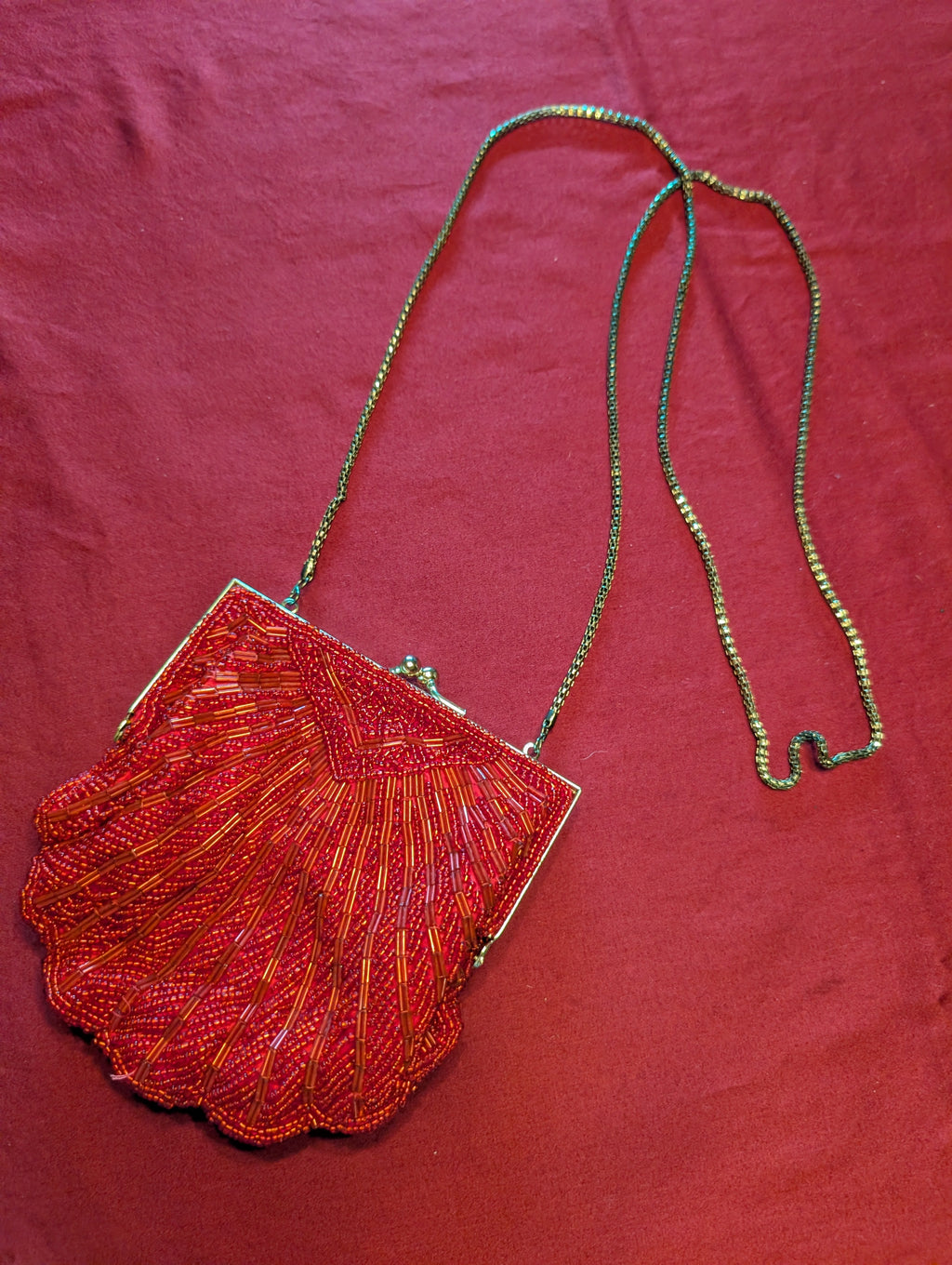 A vintage-style beaded clutch bag with a scalloped shell-shaped body, featuring red and orange beaded details, a silver-tone metal frame, and a thin metallic chain strap, displayed on a textured red fabric background.