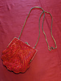 A vintage-style beaded clutch bag with a scalloped shell-shaped body, featuring red and orange beaded details, a silver-tone metal frame, and a thin metallic chain strap, displayed on a textured red fabric background.